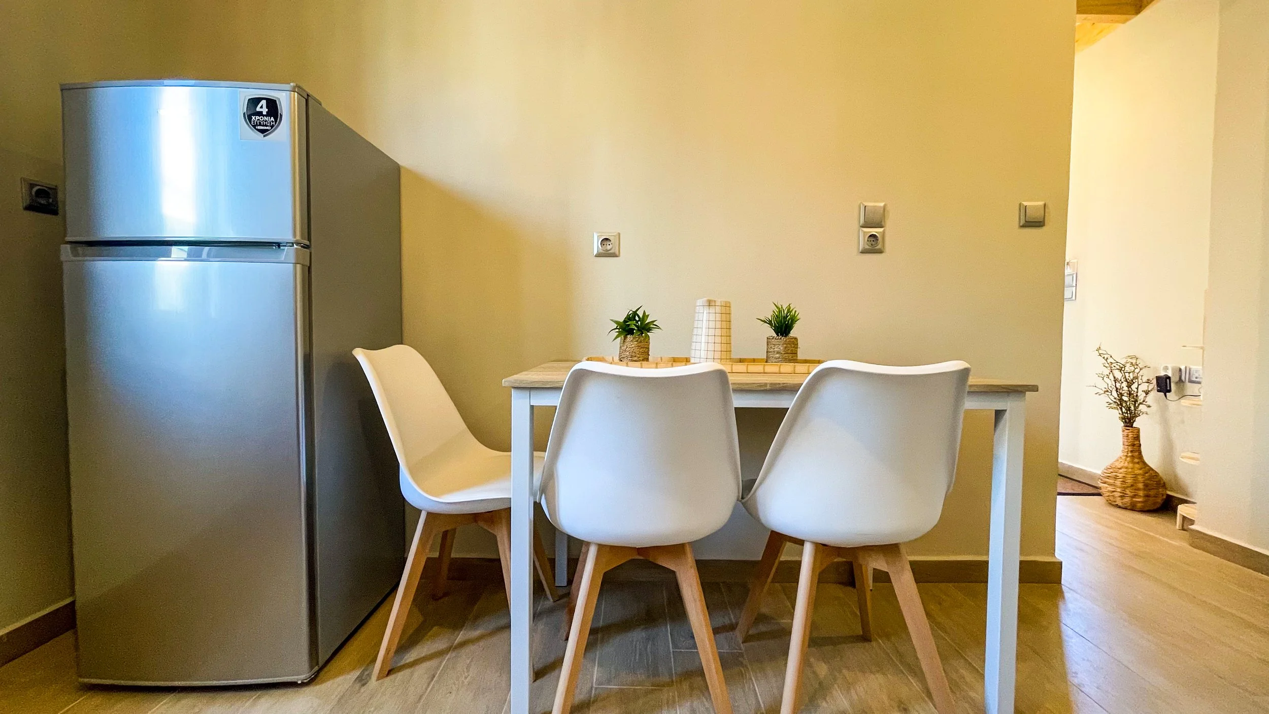 Modern dining area with a small table and three white chairs next to a stainless steel refrigerator, featuring decorative plants and a light wood floor.