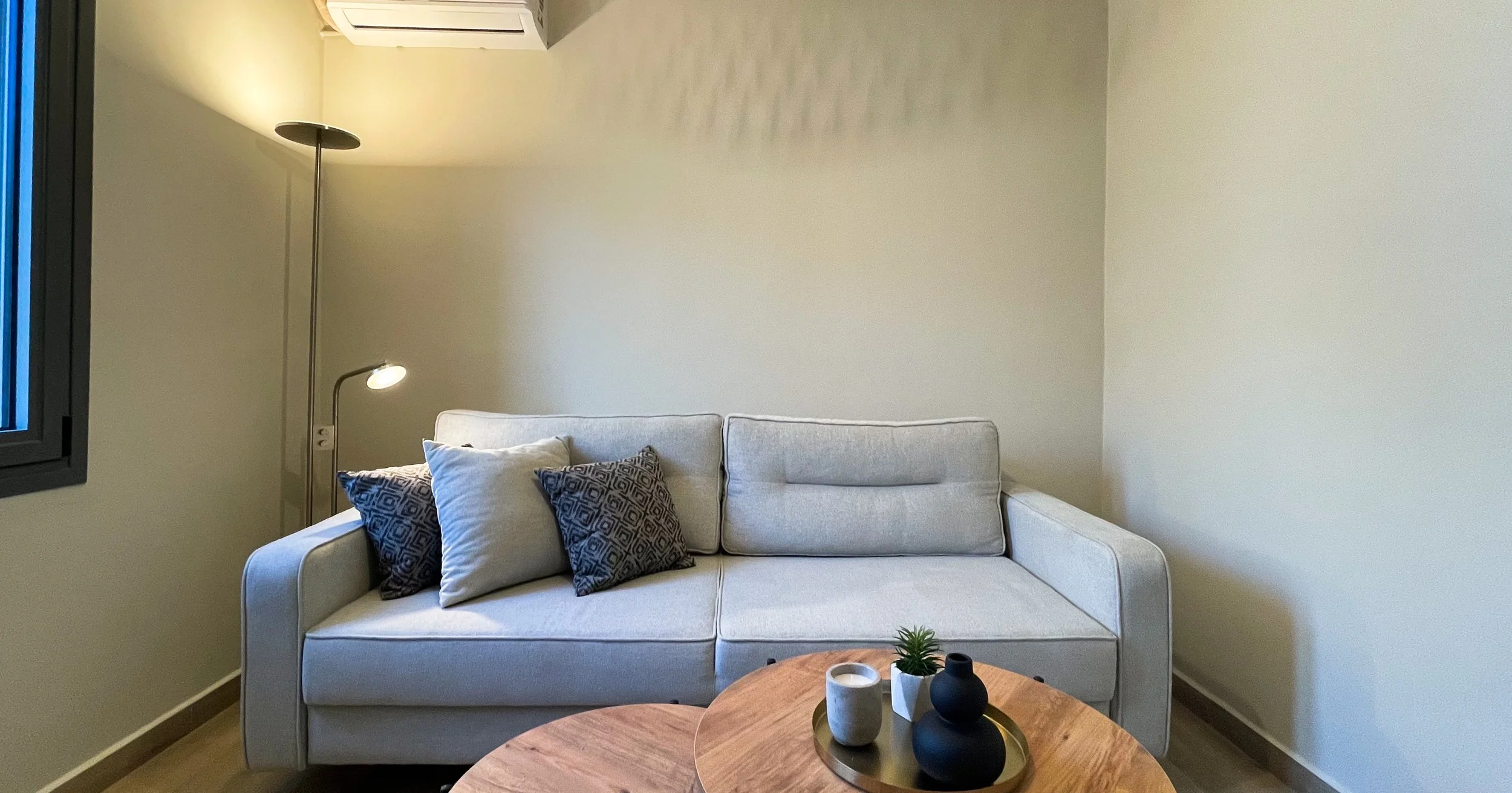 Minimalist living room with a beige sofa, decorative pillows, a floor lamp, nested wooden coffee tables, and small decorative items.