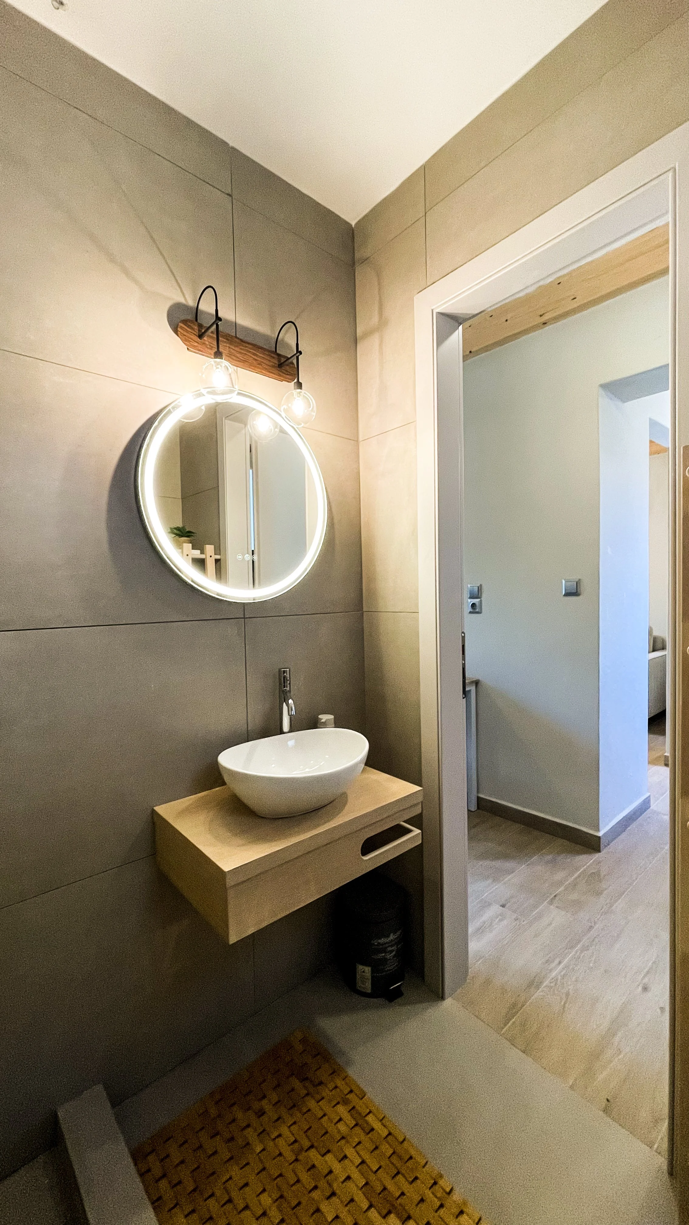 Modern bathroom with a round illuminated mirror, vessel sink on a wooden floating vanity, and beige tiled walls.