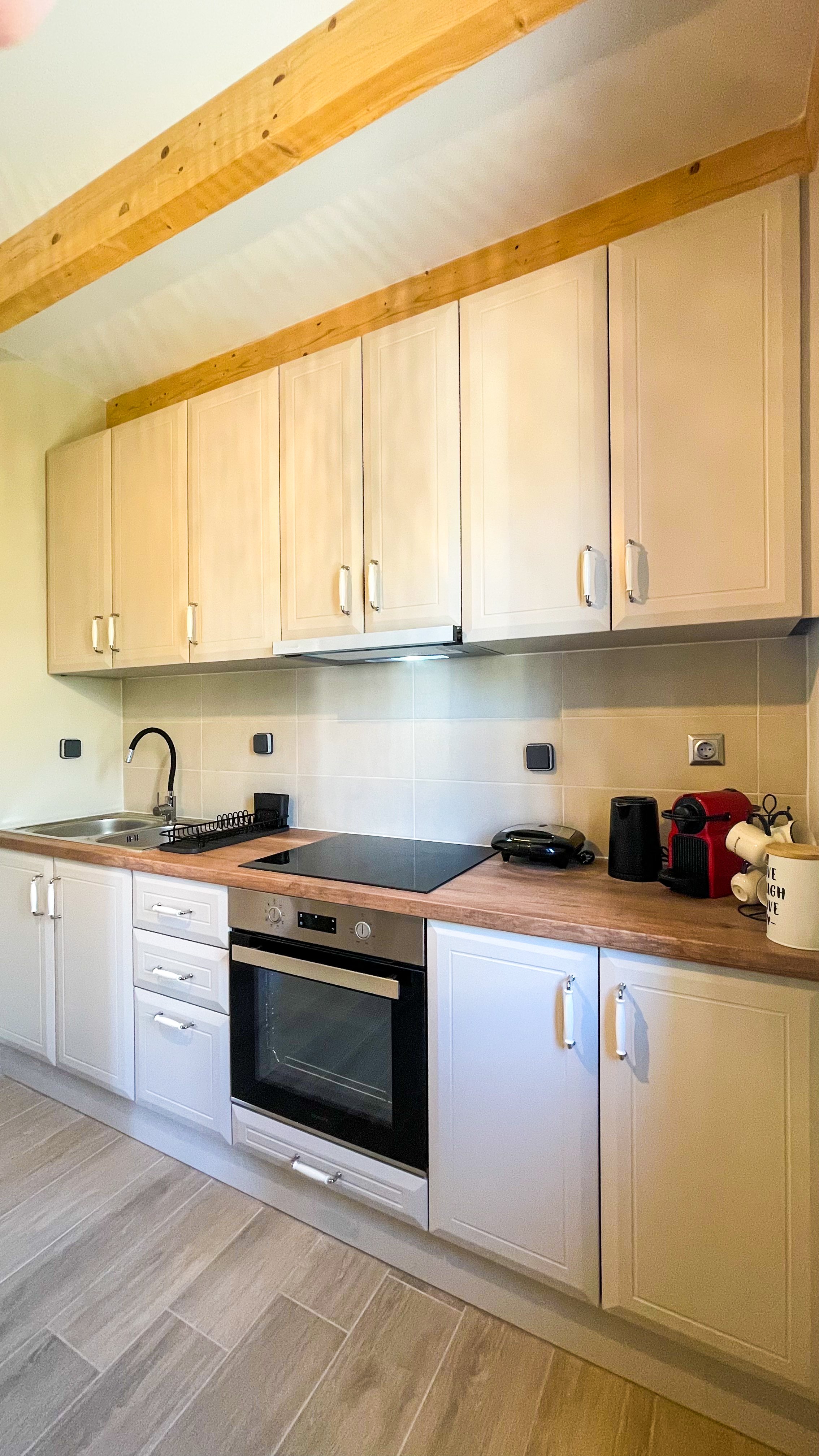 Modern kitchen with wooden cabinets, electric stove, oven, sink, coffee maker, and wooden beam ceiling. The countertop hosts a black dish rack and appliances.