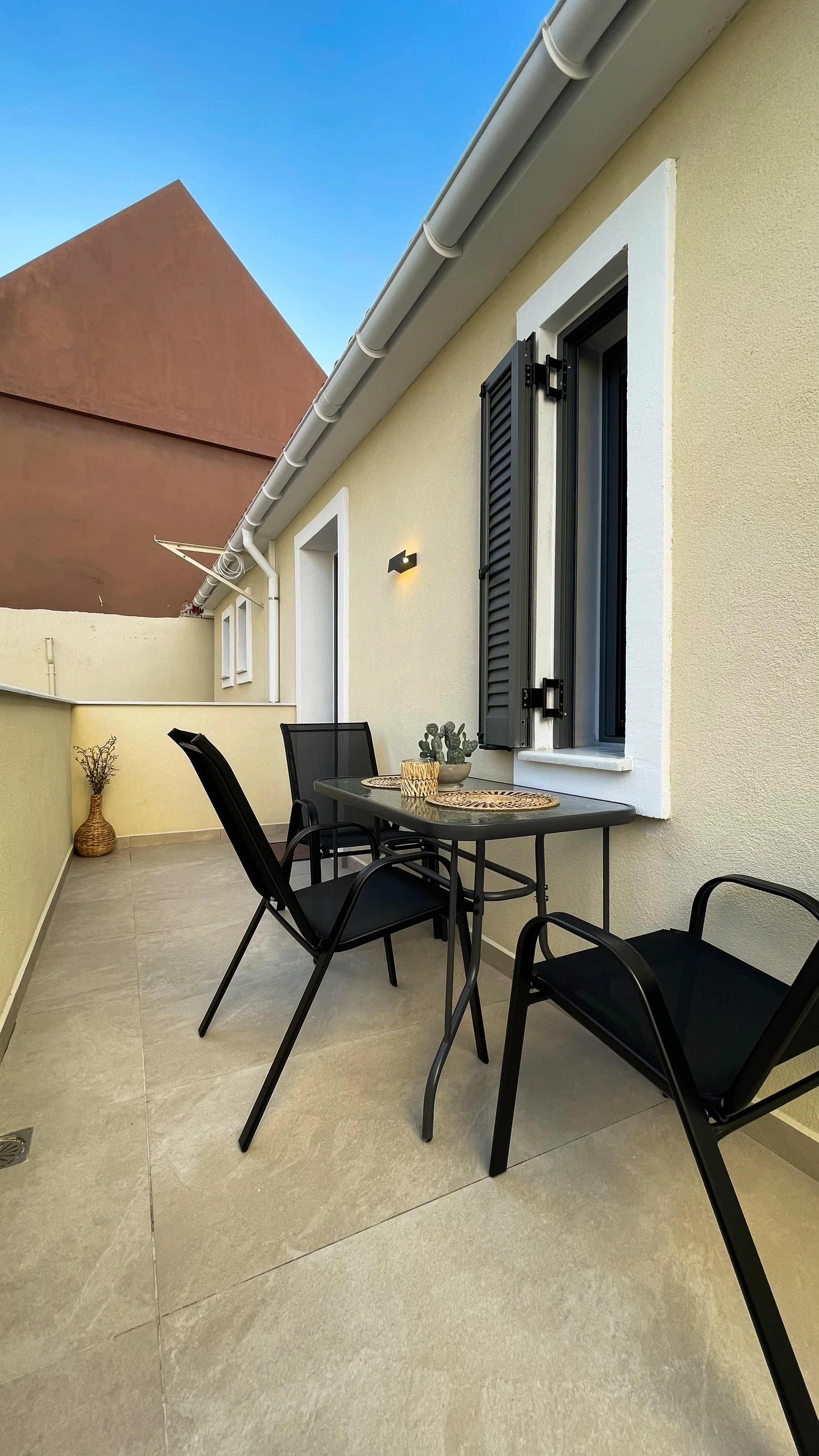 Small outdoor patio with a table and two chairs, potted plant centerpiece, and a side wall adorned with a wicker vase. Open window shutters and a wall-mounted light fixture, beige building exterior.
