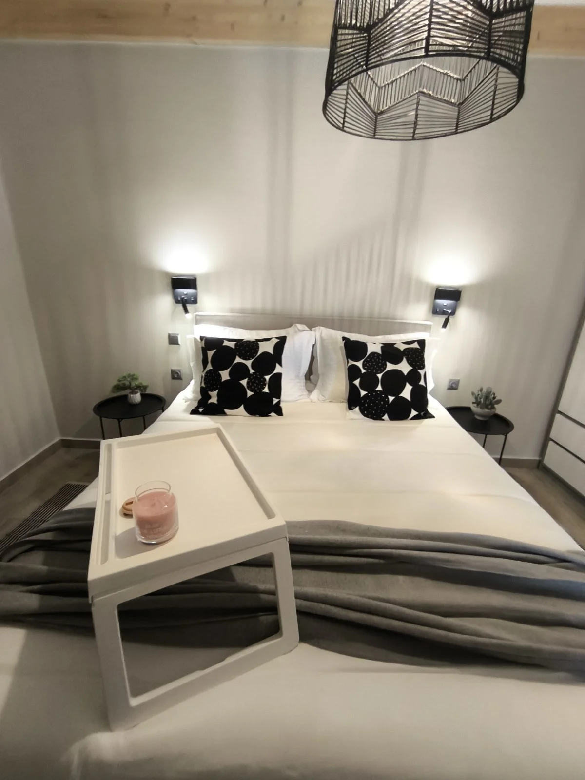 Modern bedroom with a neatly made bed featuring black and white decorative pillows and a gray throw. A small bedside table holds a plant, and a ceiling lamp hangs above. A breakfast tray with a glass of pink smoothie is on the bed.