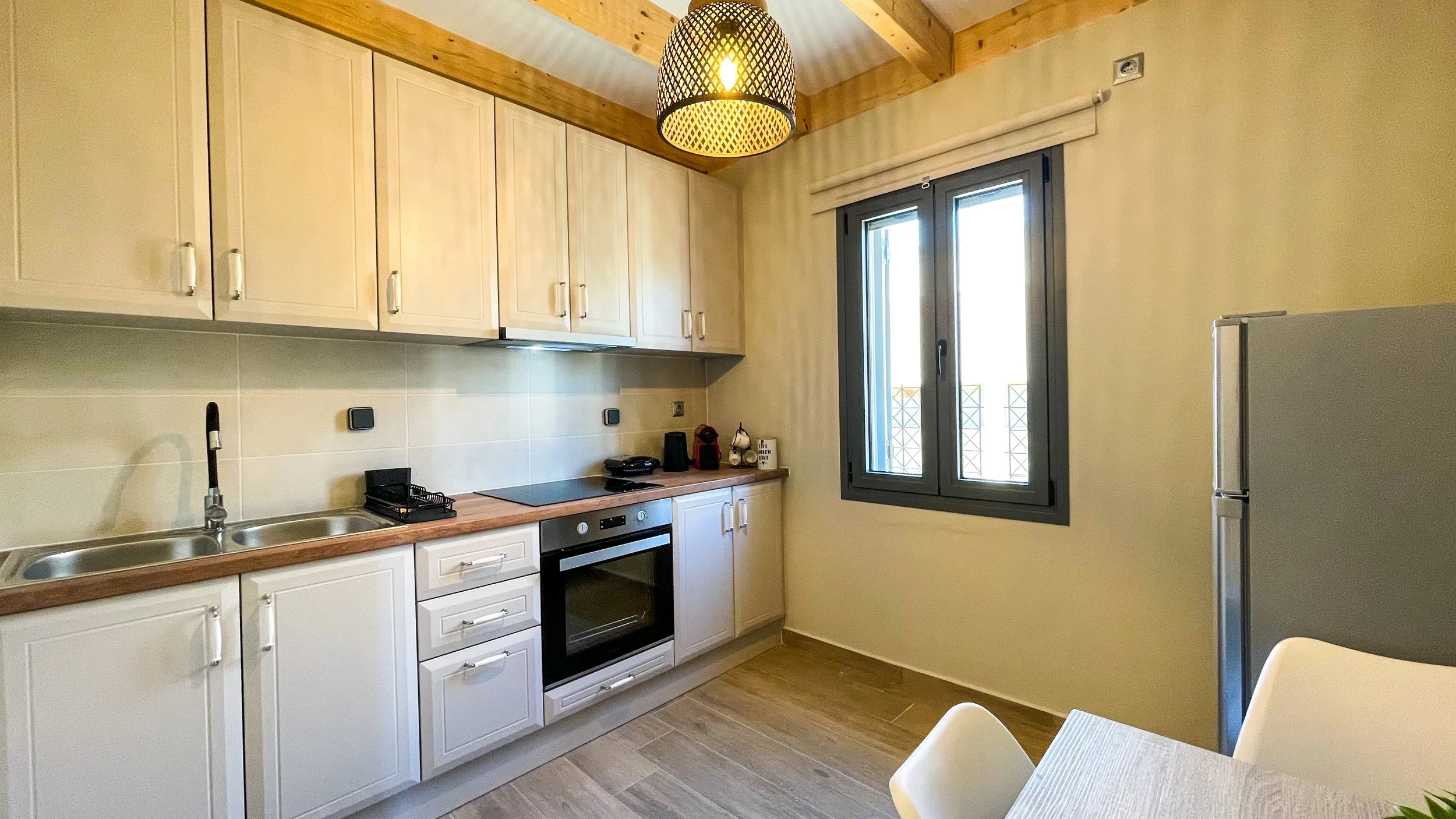 Modern kitchen with white cabinets, an oven, stove, sink, and a refrigerator, illuminated by a hanging woven light fixture. A window is present on the right side.