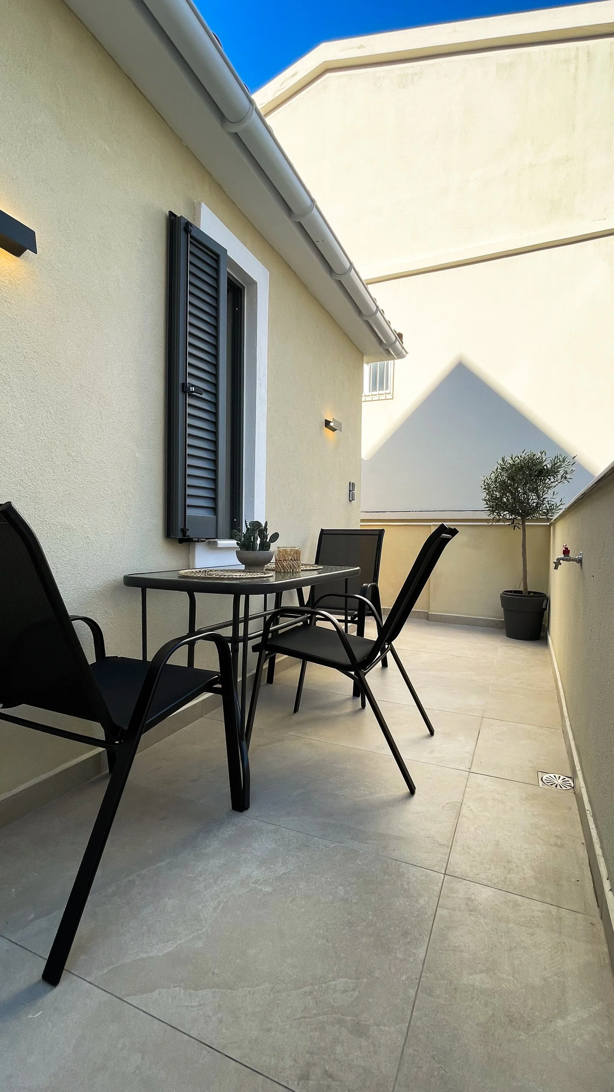 Outdoor patio with black chairs and table, potted plant, beige wall, and closed window shutters.