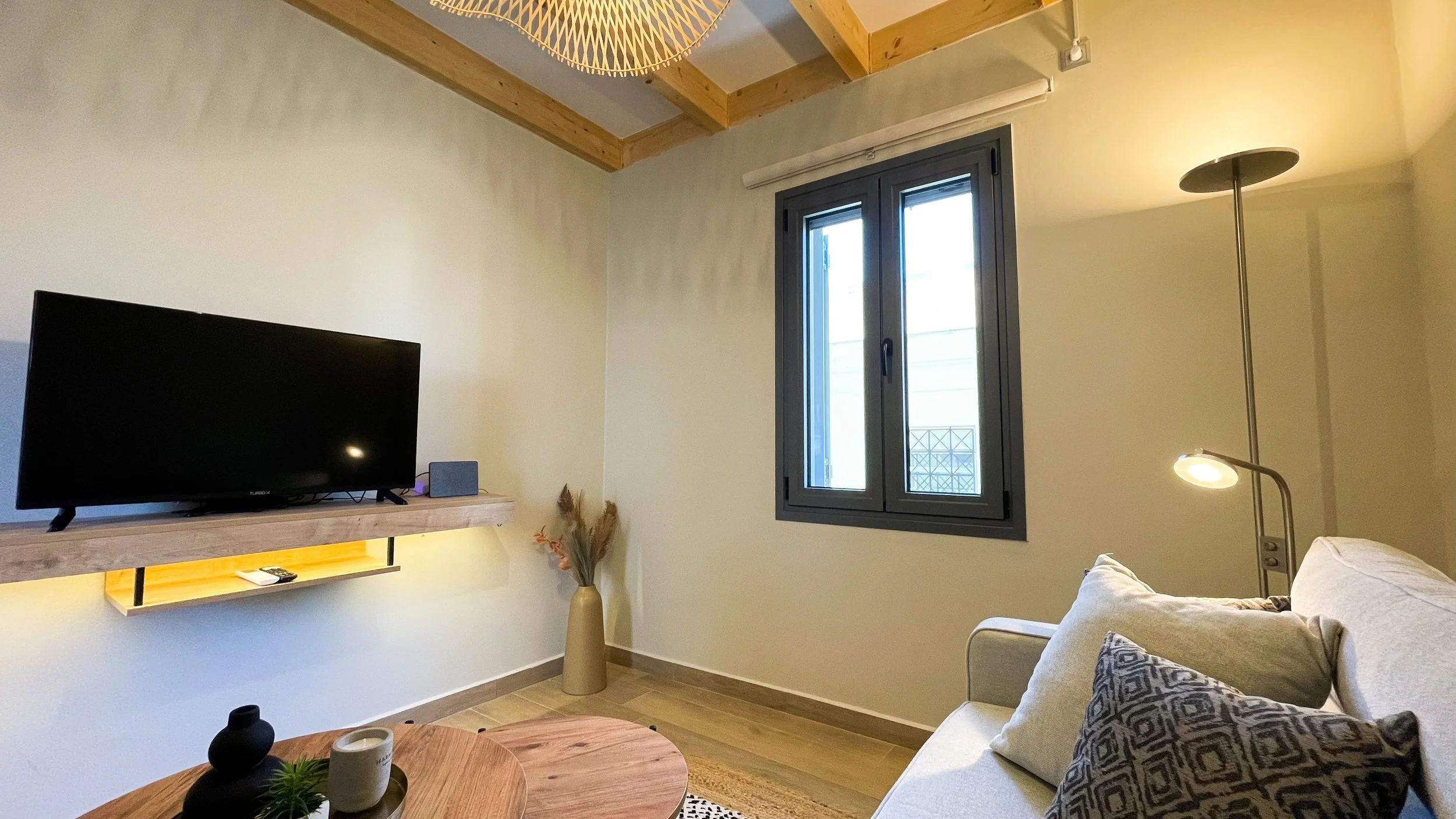 Cozy living room interior with a wall-mounted TV, beige sofa with patterned pillows, wooden coffee table, floor lamp, window, and decorative vase on hardwood floor.