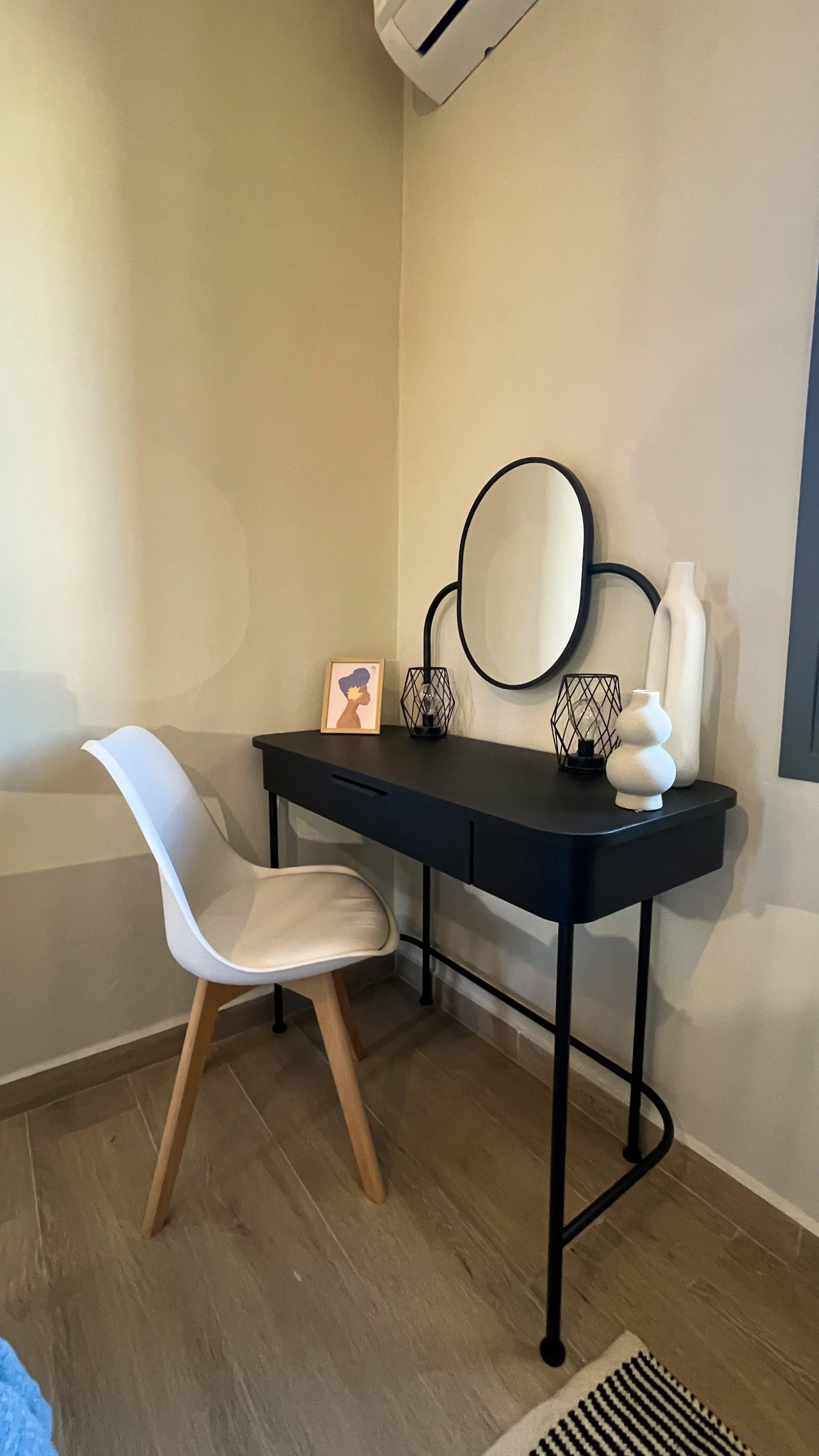 A minimalist corner of a room featuring a black desk with a round mirror, two decorative vases, and a small photo frame. Beside the desk is a white chair with wooden legs. The area has light-colored walls and wooden flooring.