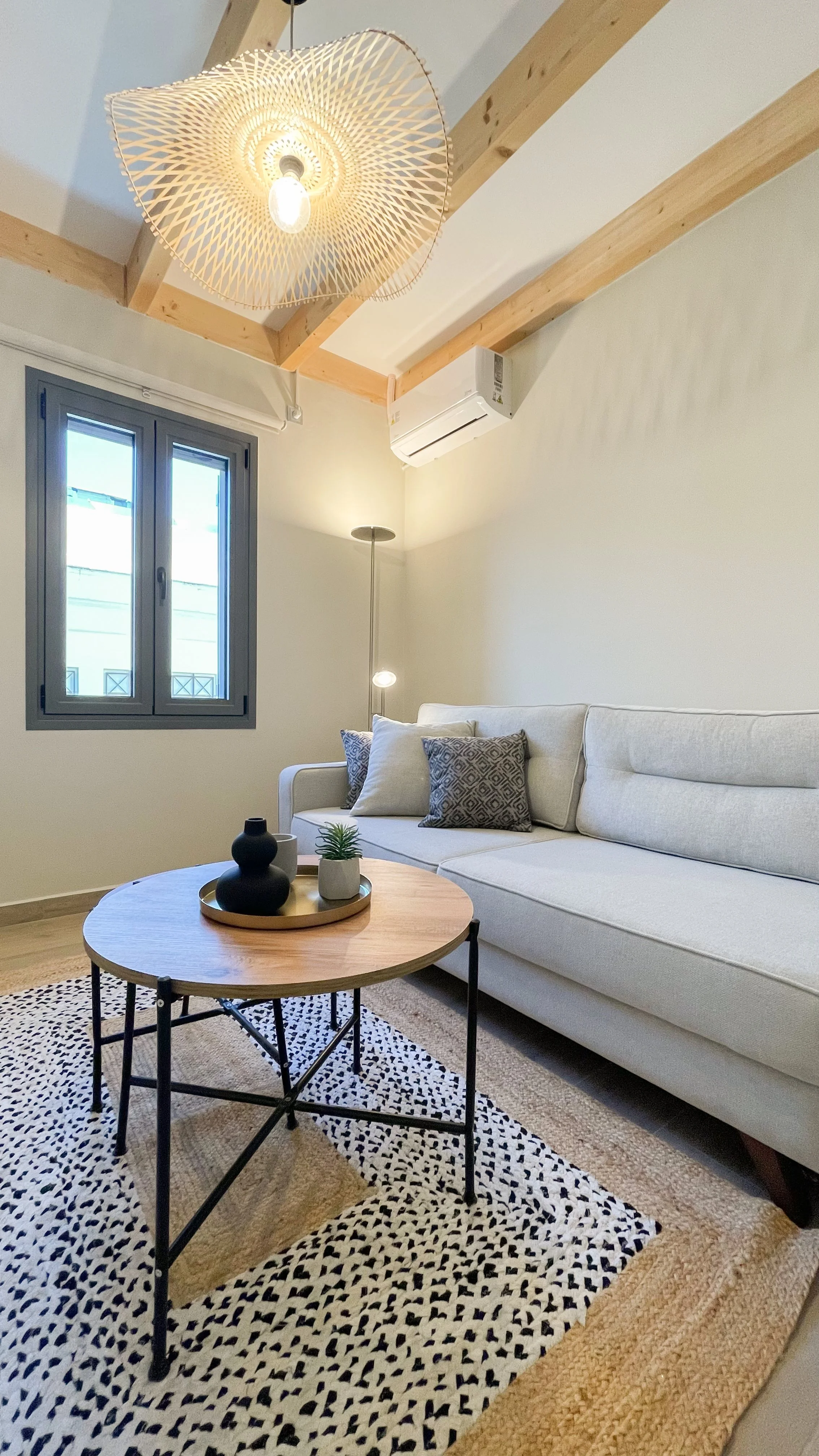 Modern living room with beige sectional sofa, decorative pillows, and a round wooden coffee table on a patterned rug. Features a ceiling light with a woven lampshade, wooden ceiling beams, and a wall-mounted air conditioner.