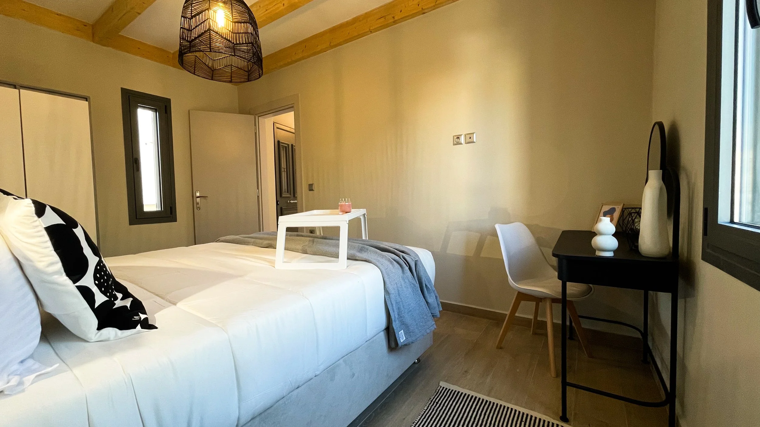 Modern bedroom with a white bed, black and white pillows, gray blanket, small desk with white chair, decorative lamp, and window lighting.