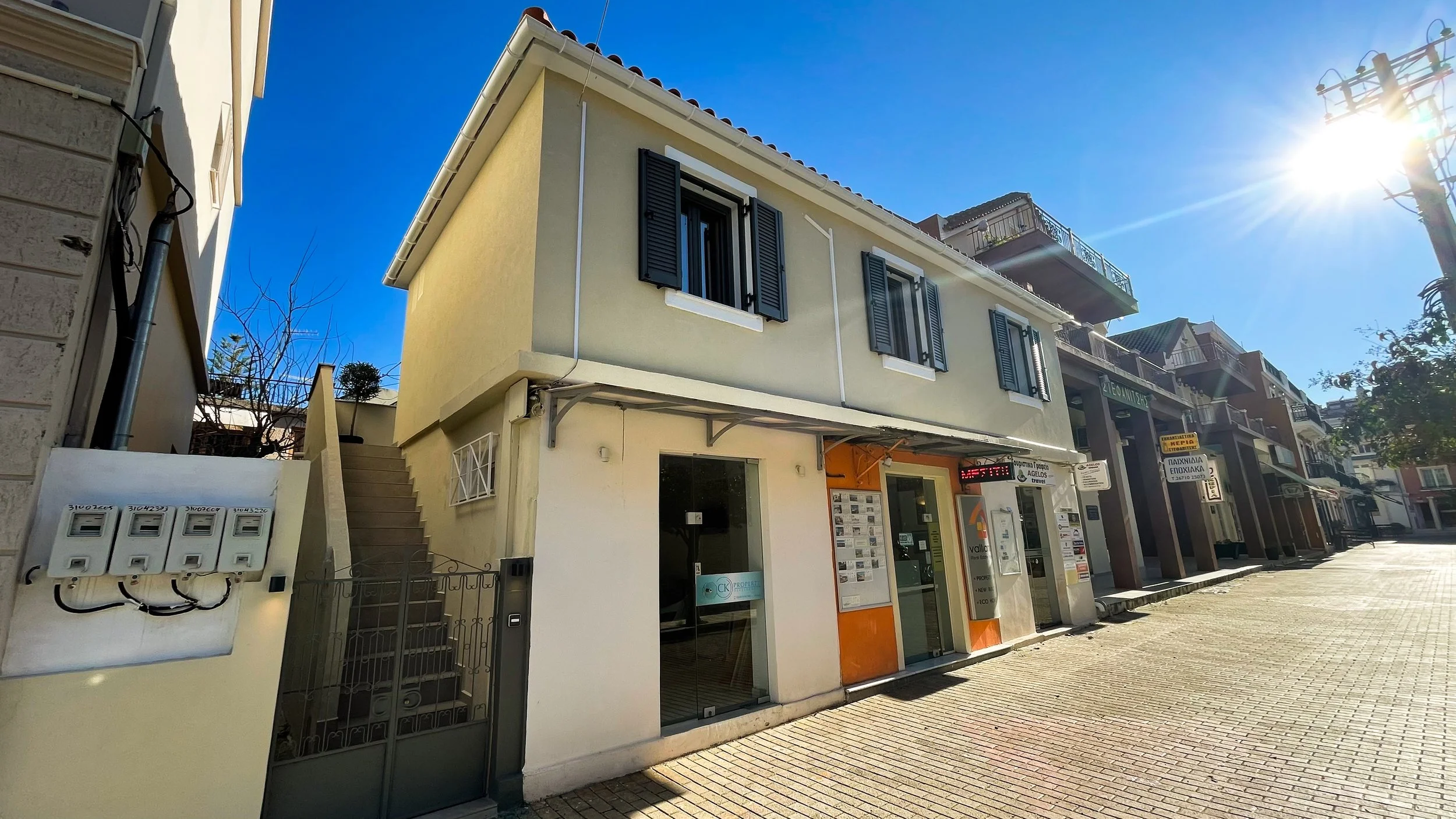 Street view of a small two-story building with beige walls, featuring a real estate office on the ground floor and shuttered windows above. Sunlight brightly illuminates the scene, casting shadows on the pavement. Additional shops stretch down the st