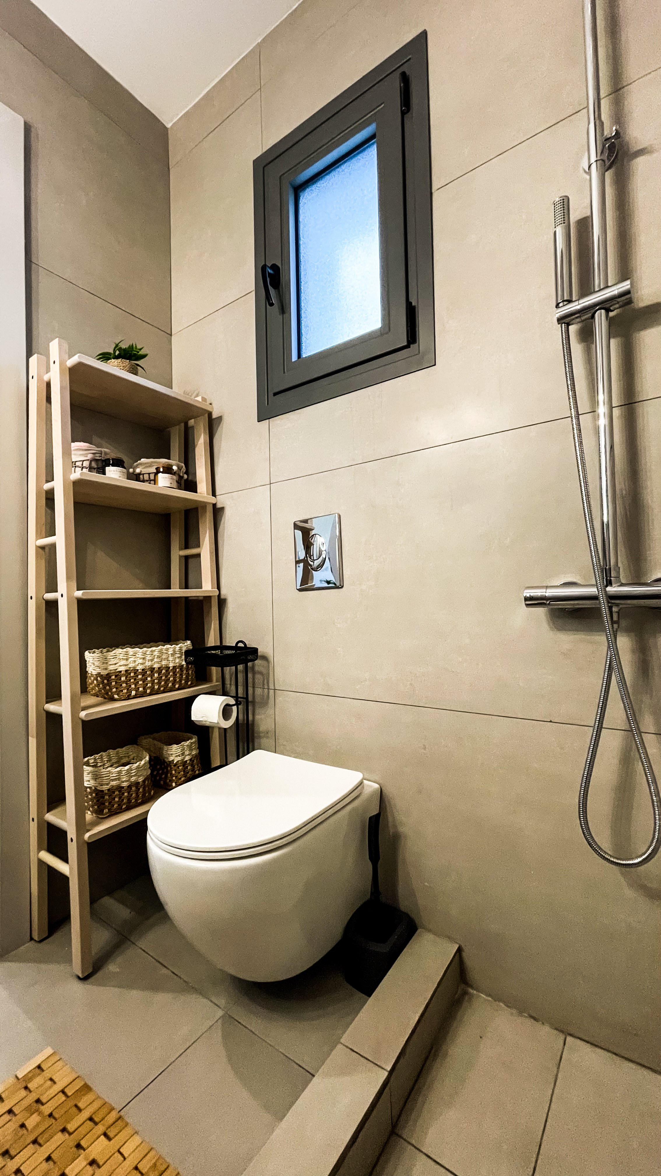 Modern bathroom with wall-mounted toilet, ladder-style wooden shelf with baskets, small window, and a wall-mounted showerhead.