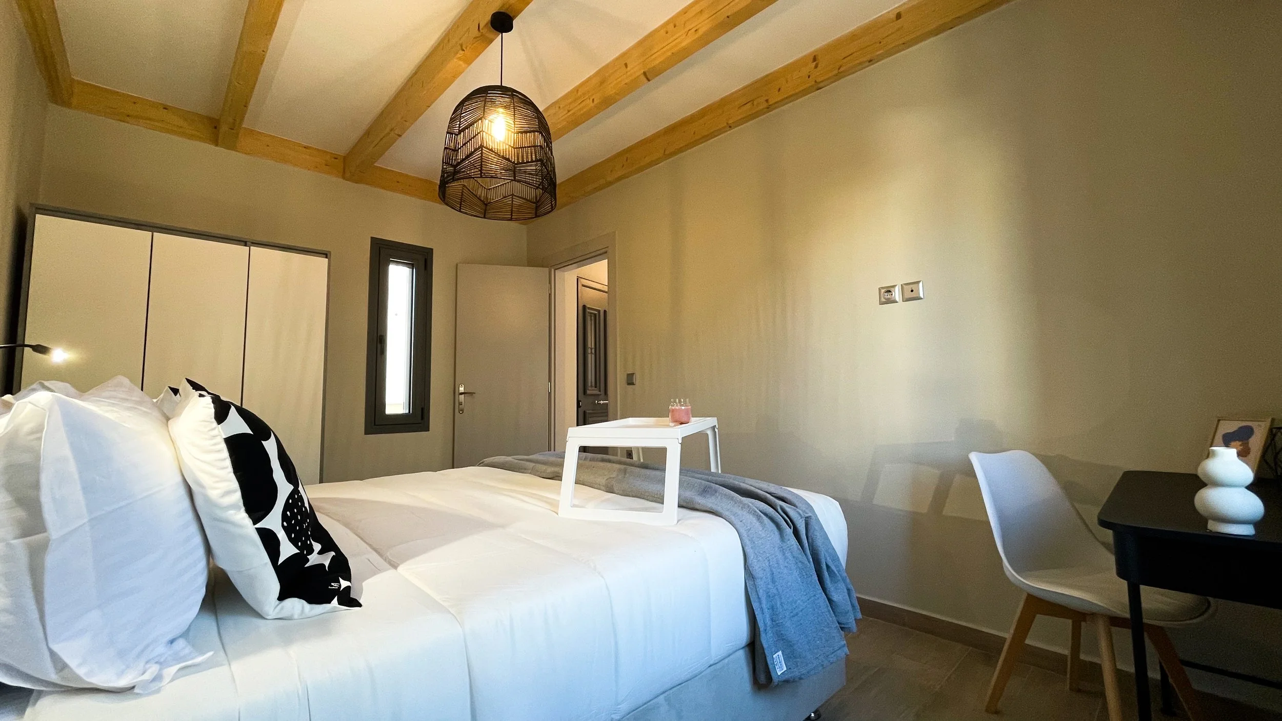 A modern bedroom with exposed wooden beams, featuring a comfortable white bed with black and white accent pillows and a folded gray blanket. A white tray holds a small candle. To the right, there's a black desk with a white chair, decorated with a sm
