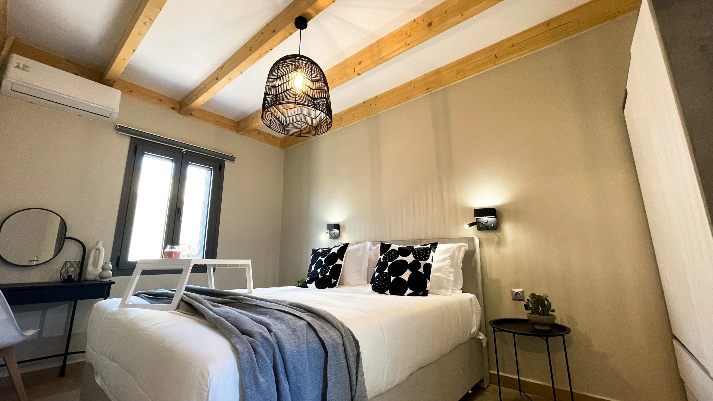 Modern bedroom with wooden beams, a large double bed with black and white pillows, gray blanket, and a white tray. A round mirror and desk are to the side, with a window above. A hanging lamp and small plant add decor. Air conditioner above the windo