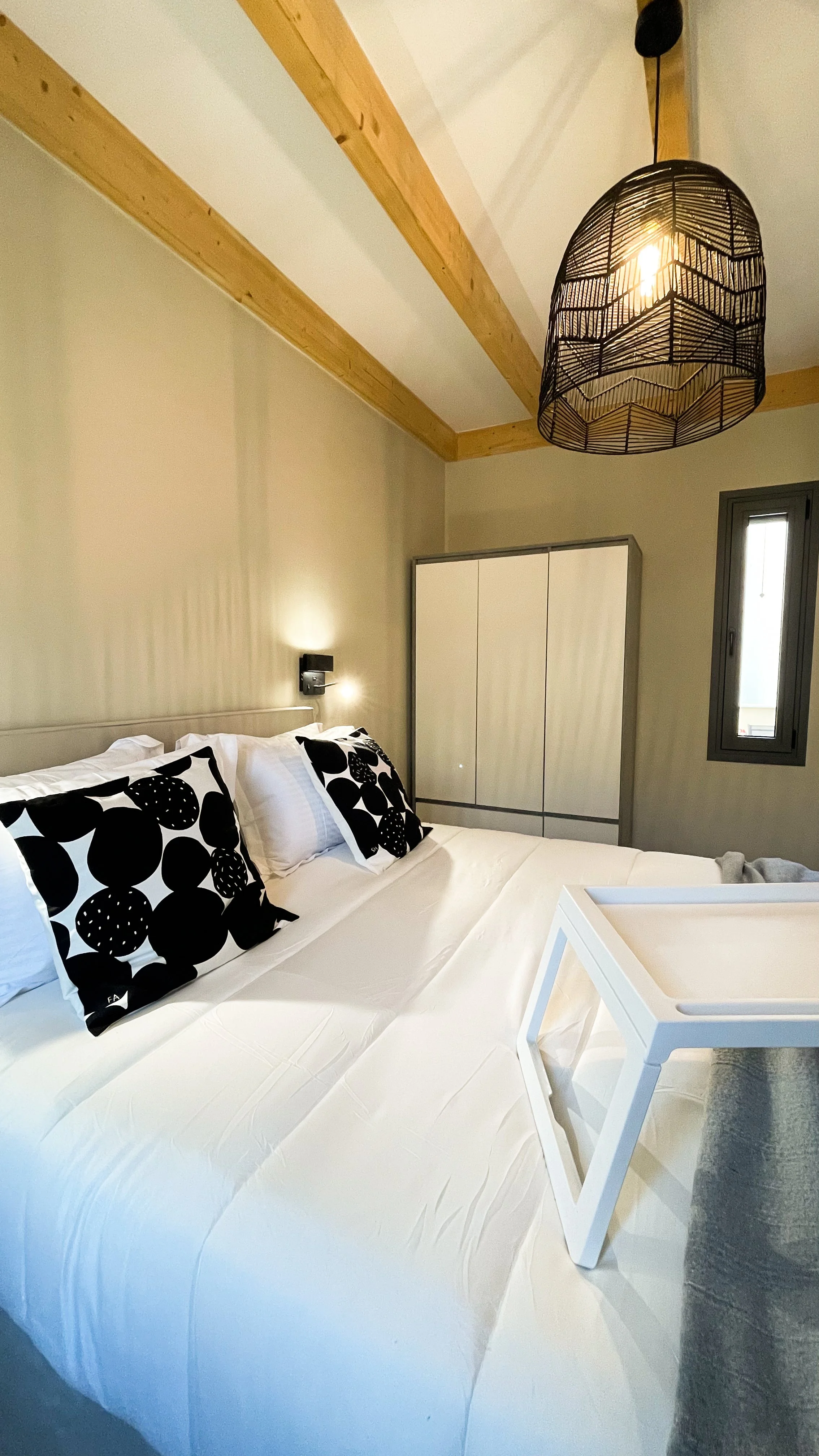 Modern bedroom with a white bed, black and white geometric pillows, wooden ceiling beams, pendant light, white furniture, and a small window.