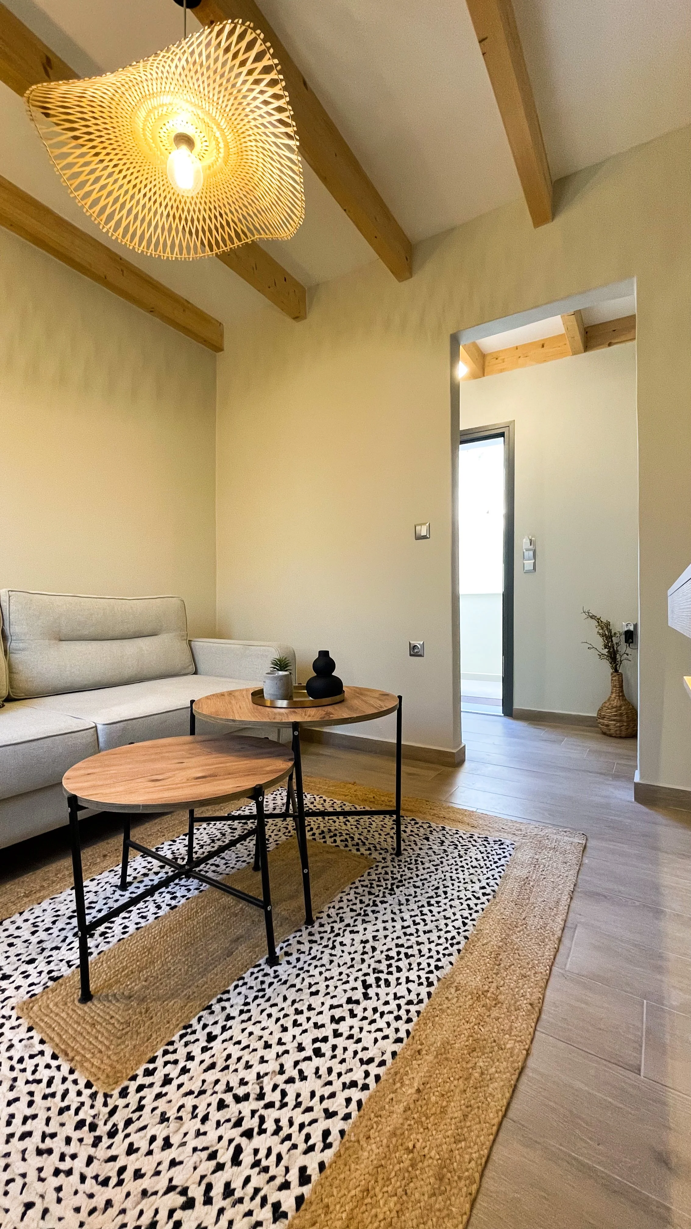 Modern living room with a beige sofa, two wooden coffee tables on a patterned rug, a woven pendant light, and visible wooden ceiling beams. A decorative vase and plant are on the tables.