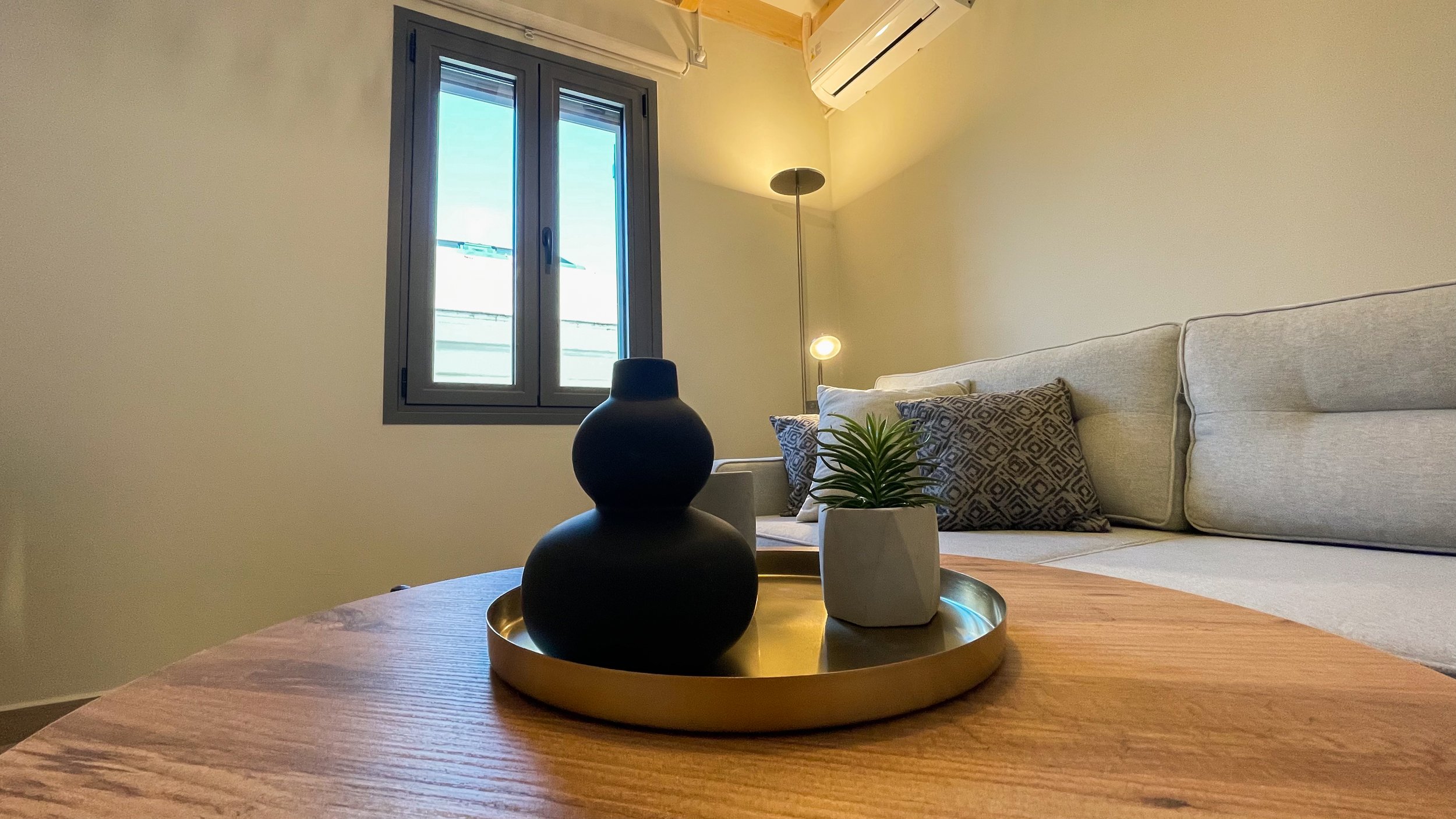 Modern living room with a wooden coffee table, featuring a black vase and a small potted plant on a tray, a gray sofa with patterned cushions, a window, and a floor lamp.