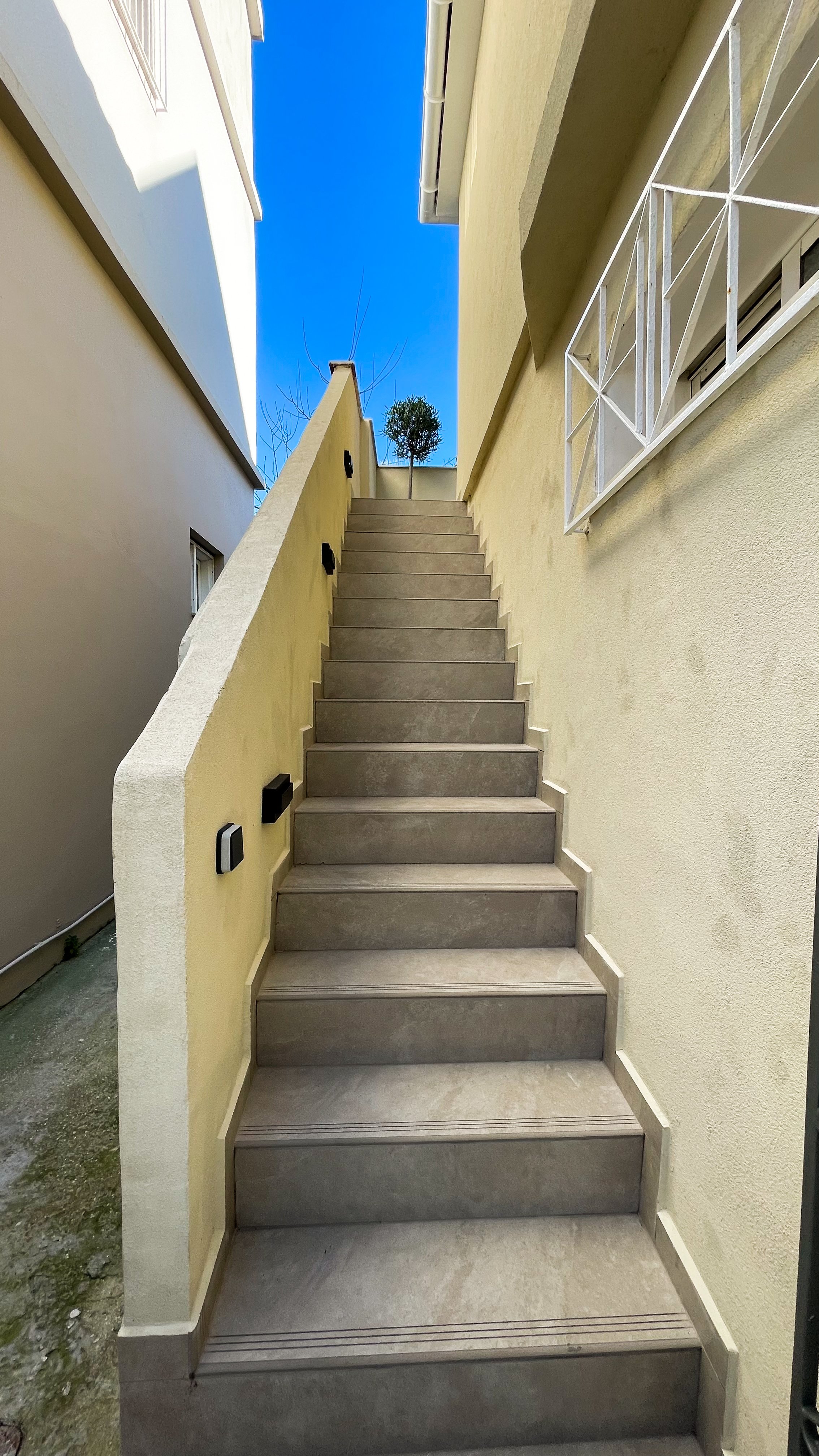 Outdoor stone staircase with beige walls, leading to a tree at the top, under a clear blue sky, featuring white railing on the right side.