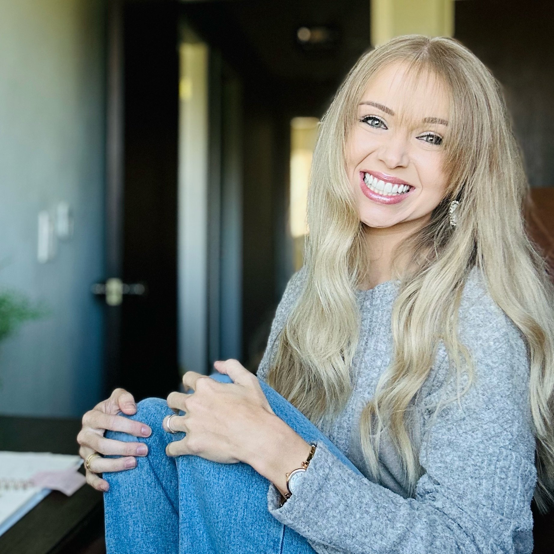 Smiling woman with long blonde hair in gray sweater and blue jeans representing approachable, client-focused service.