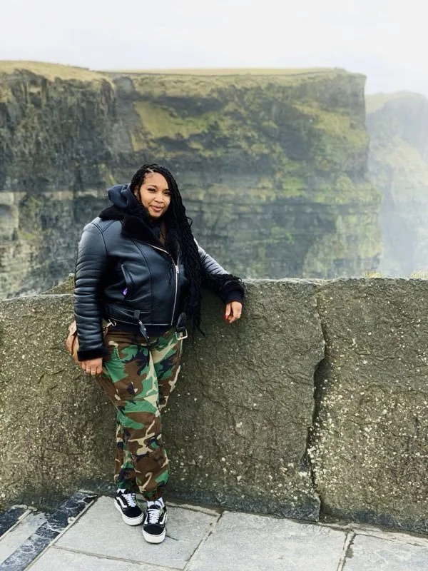 A woman stands next to a stone railing at the edge of a cliff in front of a scenic view of green cliffs and foggy sky.