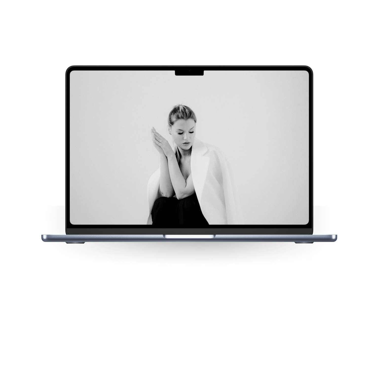 Black and white photo of a woman on a laptop screen, with her hair in a bun, wearing a white jacket over a black outfit, with her hand near her head, looking thoughtful.