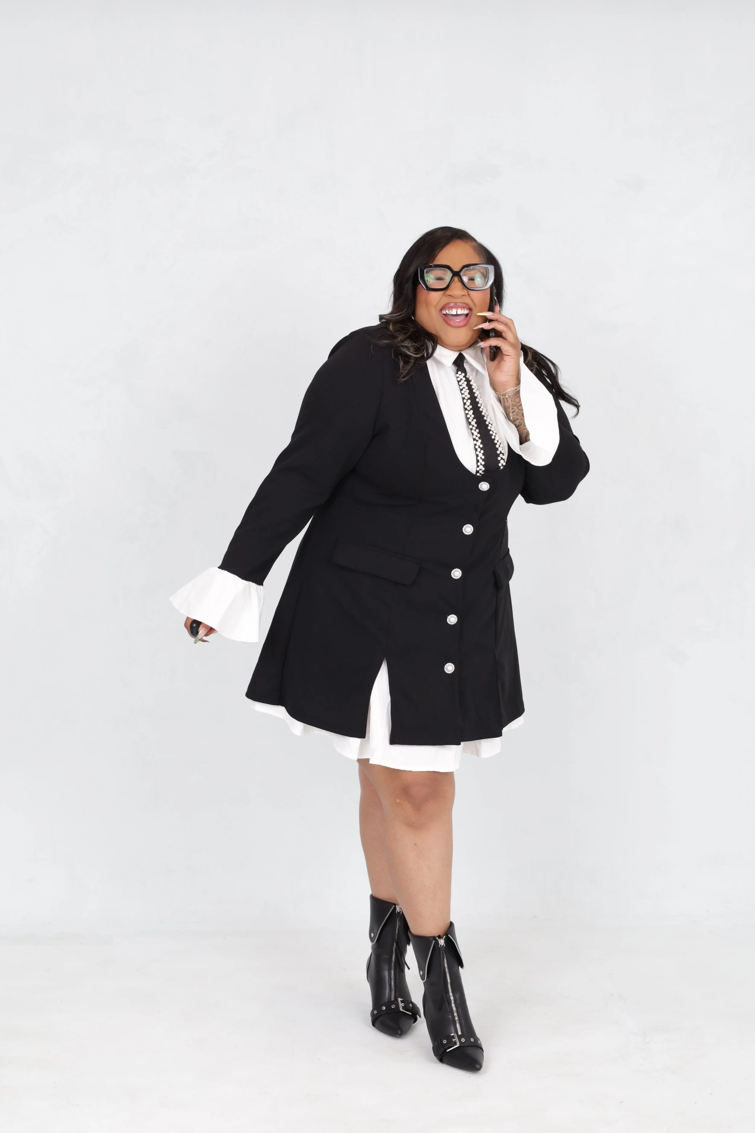 A woman with glasses talking on a cellphone, wearing a black blazer over a white dress with bell sleeves, standing against a plain white background.
