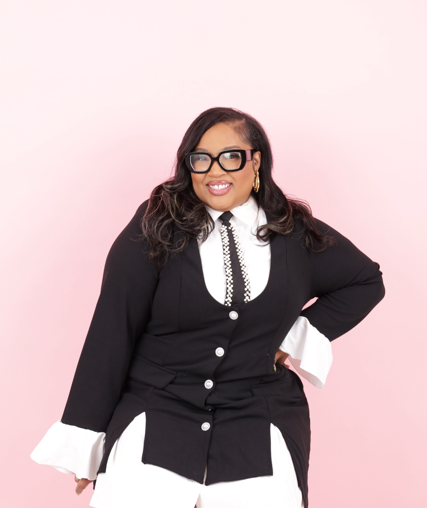 Woman with long dark hair, wearing large black glasses, gold hoop earrings, a black blazer, and white blouse with a pearl necklace, standing against a pastel pink background.