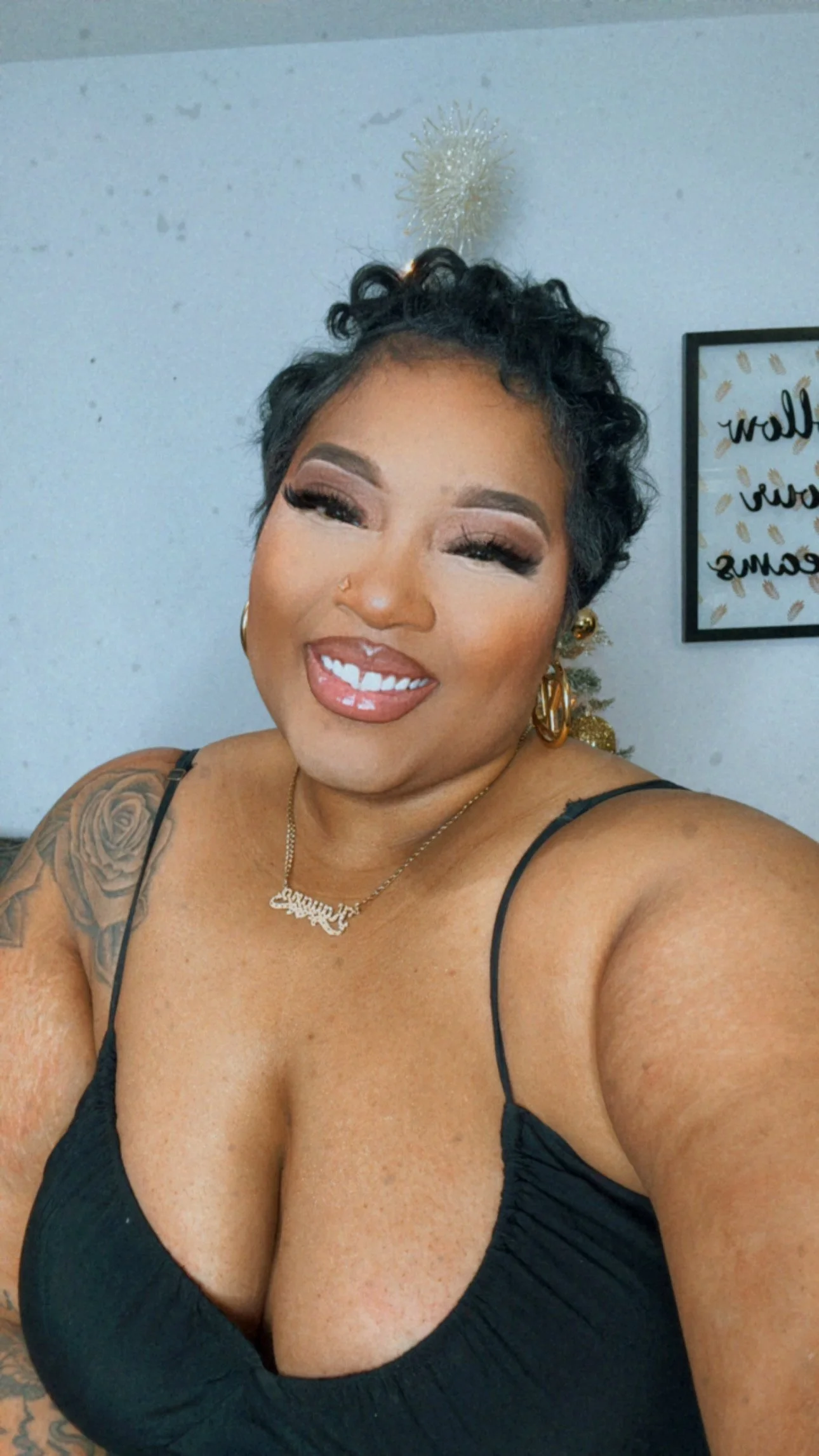 A woman smiling, with short curly hair, wearing makeup, gold earrings, a nose ring, and a gold necklace, dressed in a black spaghetti strap top with a tattoo on her left shoulder, standing in front of a wall with a framed sign and holiday decorations.