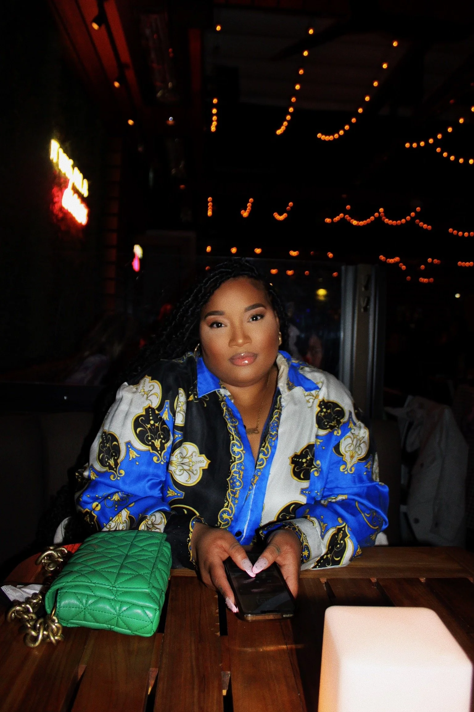 A woman with dark braided hair sitting at a wooden table in a dimly lit restaurant, gazing at the camera, with vibrant makeup and wearing a colorful patterned shirt. She has a green quilted handbag with a gold chain and clasp on the table in front of her, and her hands are holding a smartphone. The background has warm string lights hanging from the ceiling.