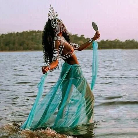 A woman in a flowing turquoise skirt and tribal accessories stands in a body of water, holding a mirror and a piece of fabric, with trees in the background.