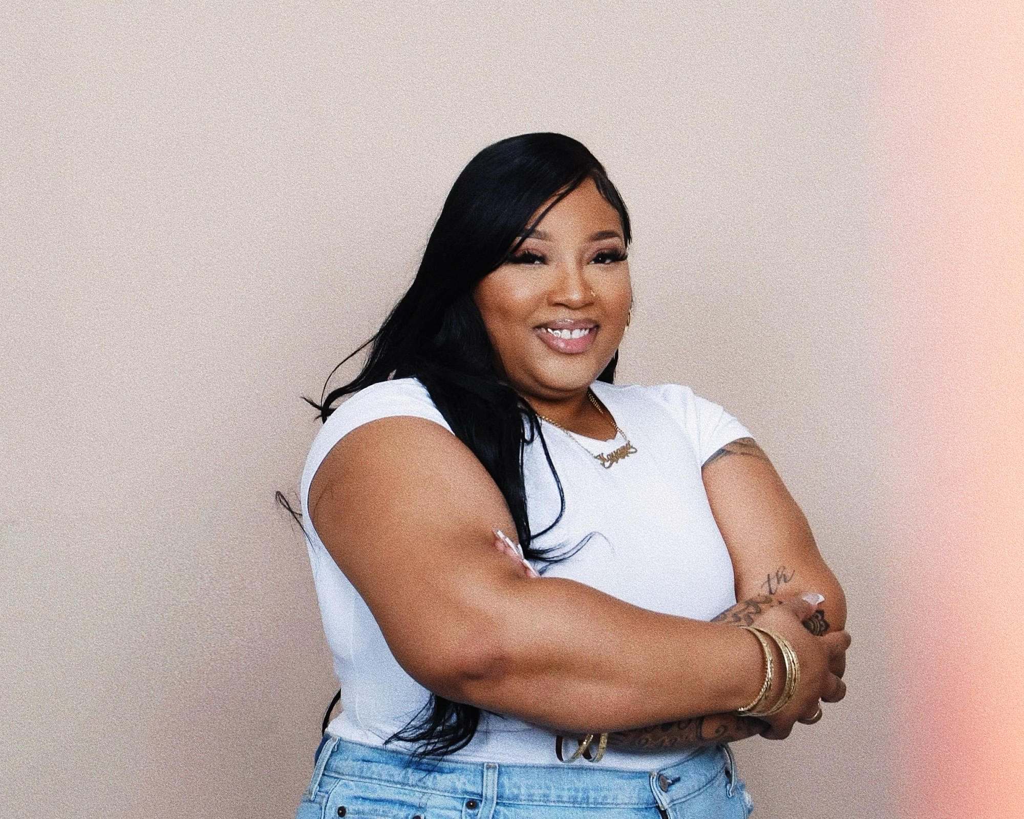 A woman with long black hair wearing a white t-shirt and blue jeans, smiling and crossing her arms, standing against a plain wall background.