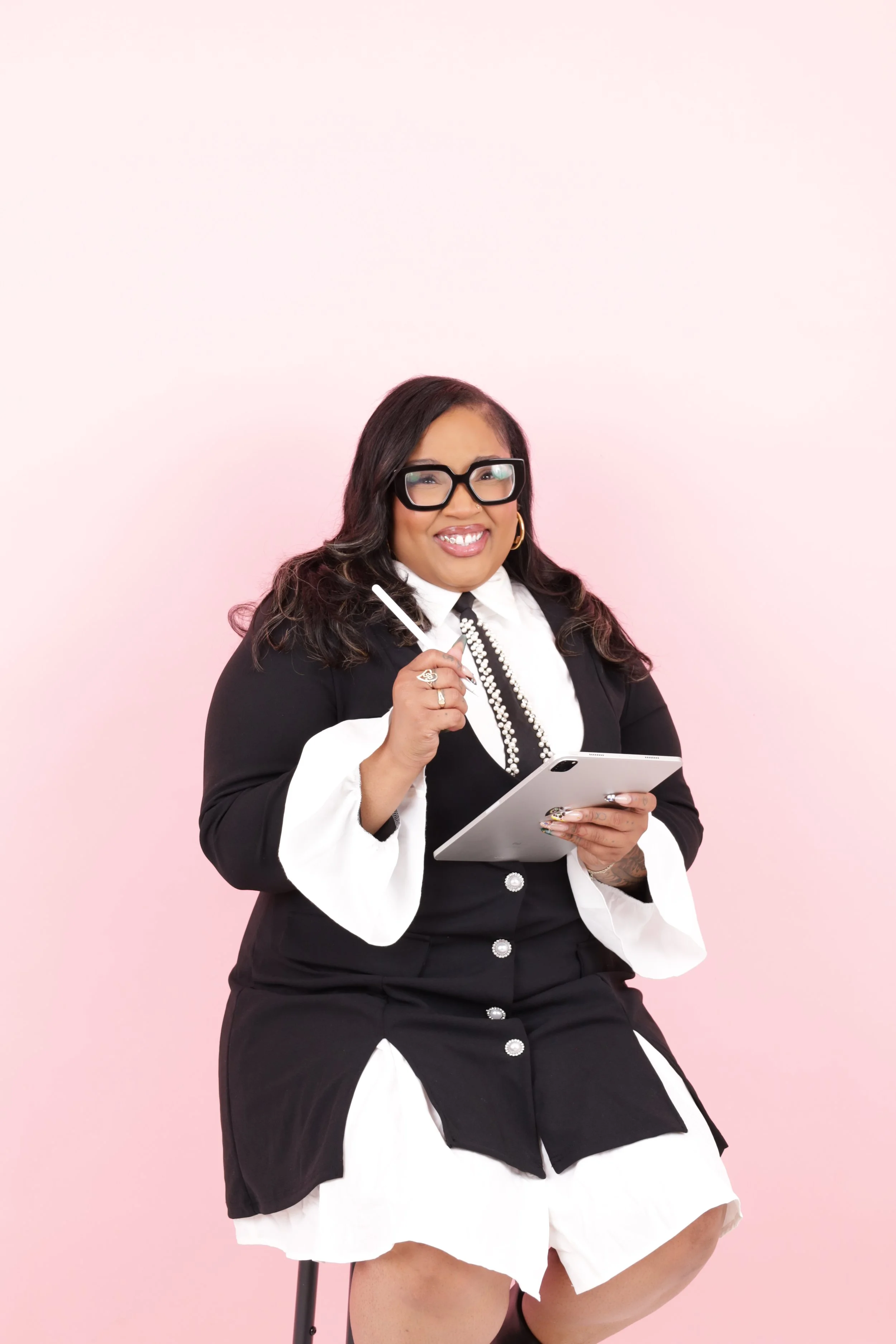 A woman sitting in front of a pink background, wearing glasses and a black and white outfit, holding a tablet and a stylus, smiling.