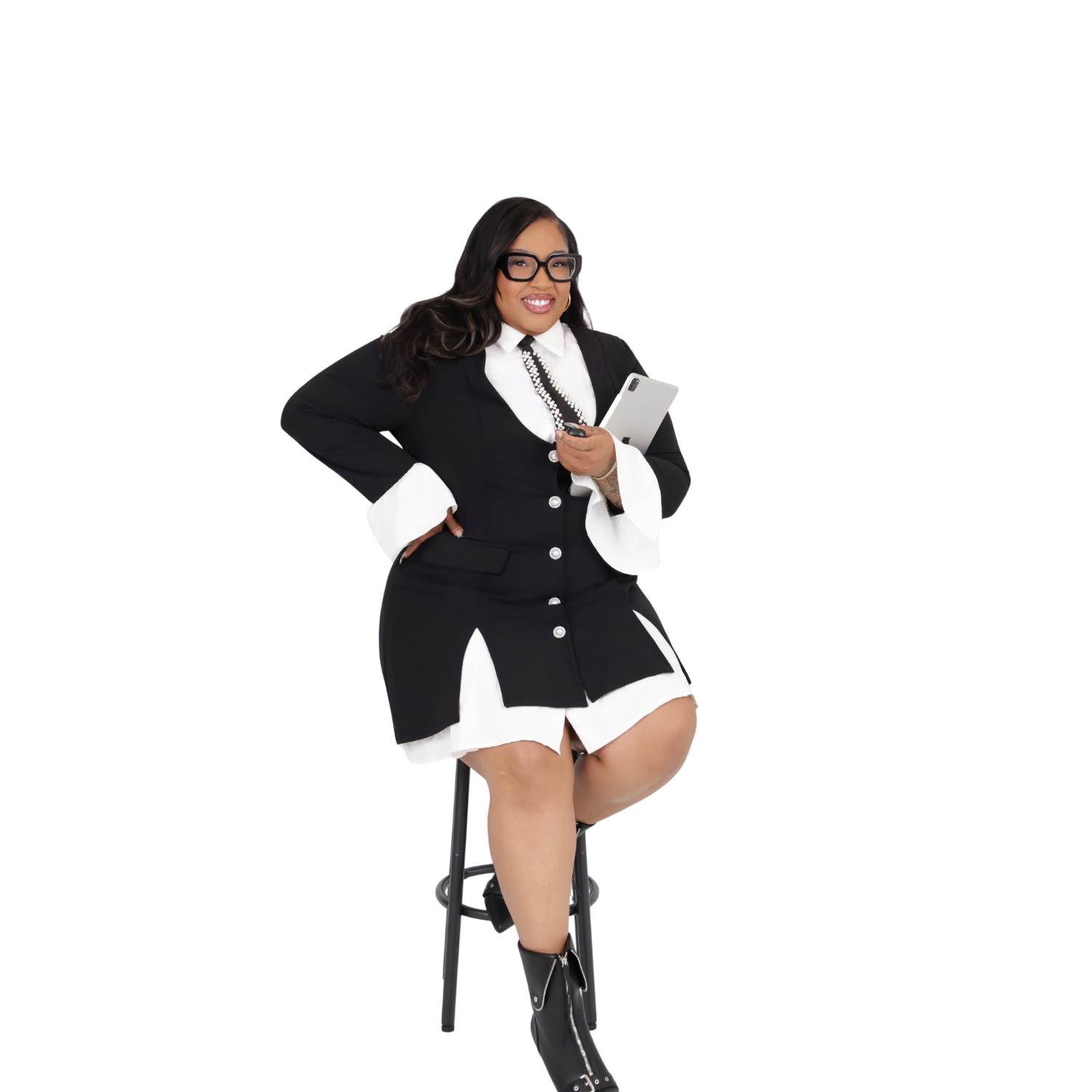 A woman dressed in a black and white suit, wearing glasses, holding a tablet, sitting on a stool, smiling.
