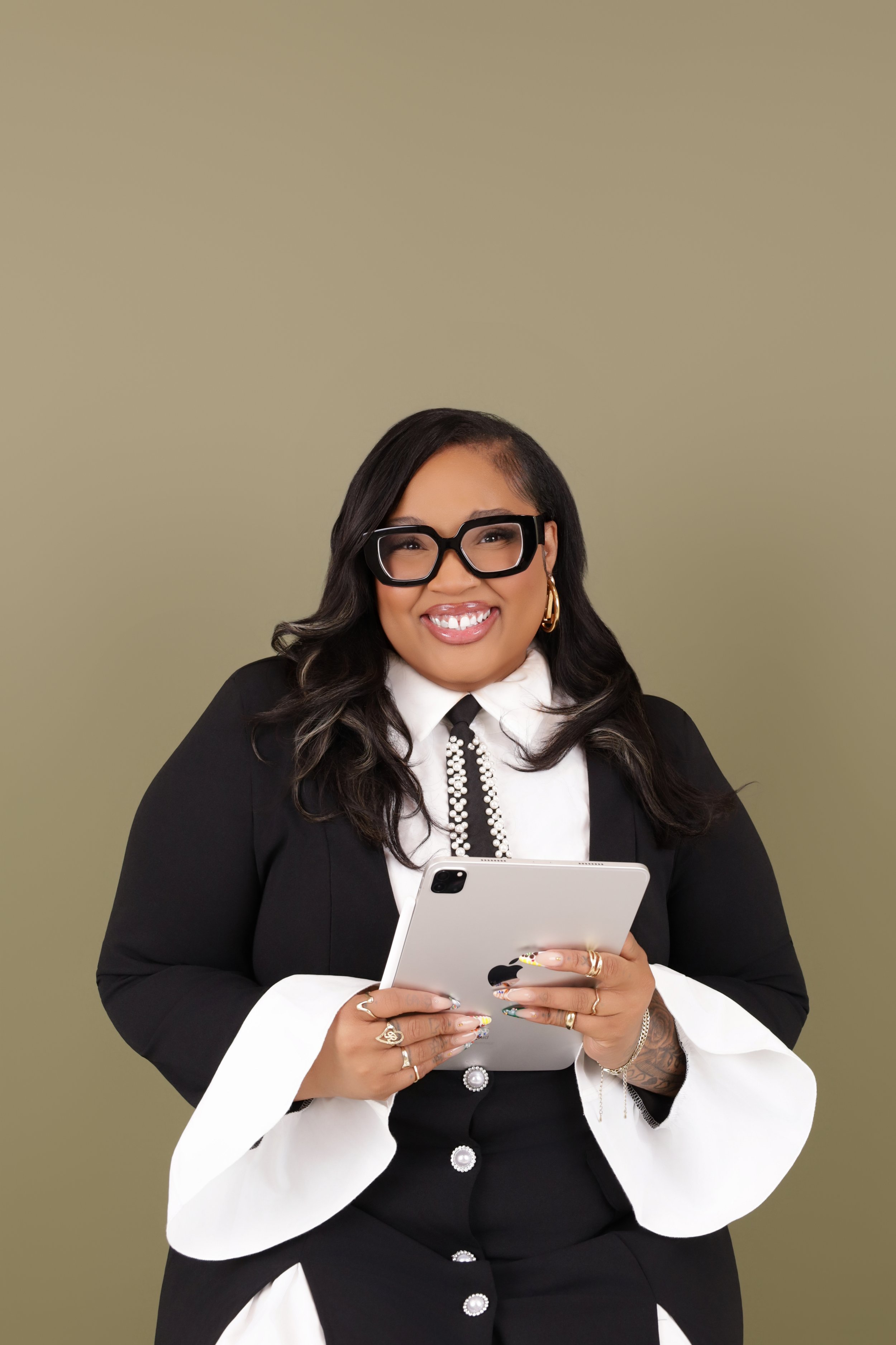 Smiling woman with dark hair, glasses, and jewelry holding a tablet against a plain background.