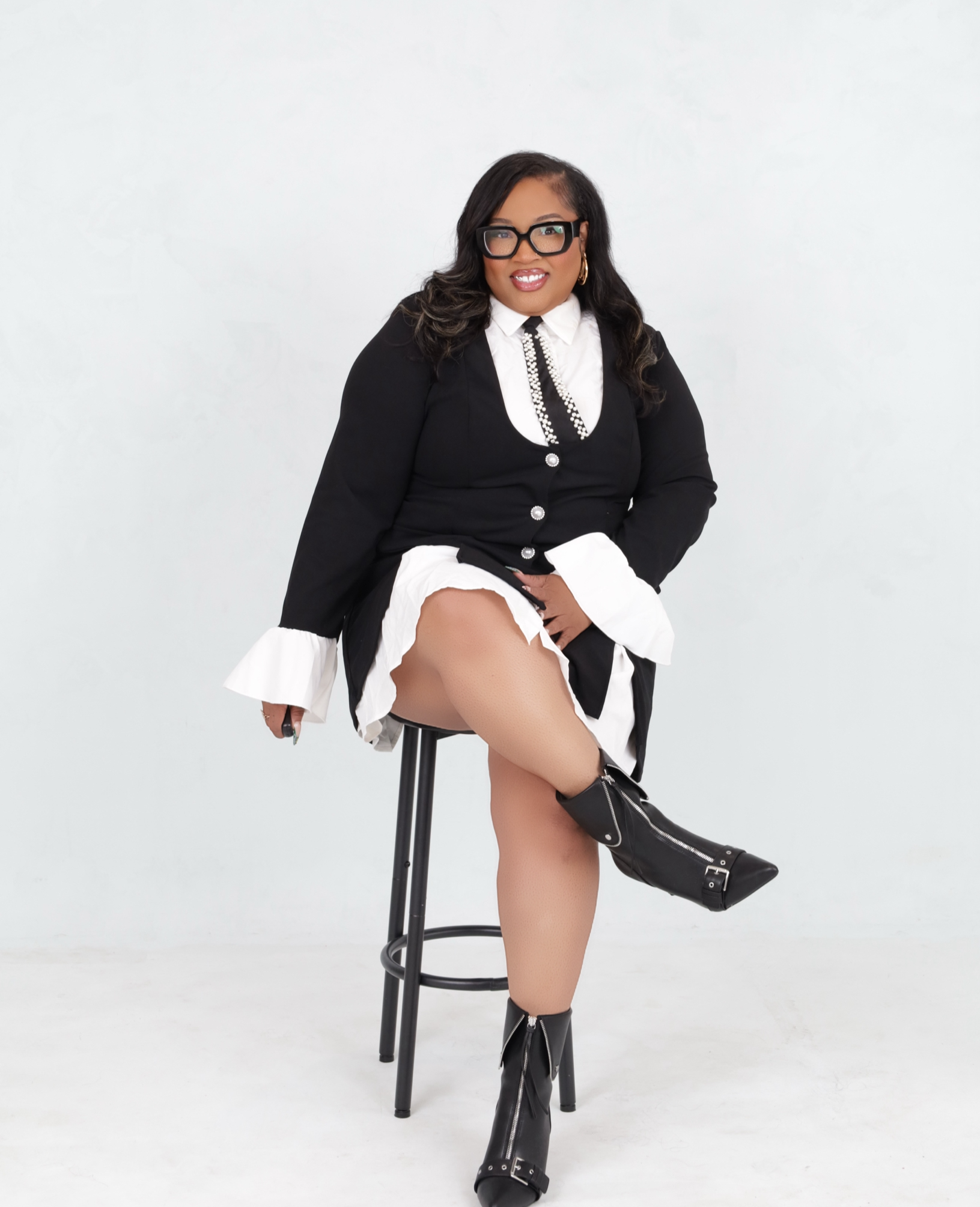 Woman sitting on a stool, wearing black and white outfit with black boots, posing with her leg crossed, against a plain white background.