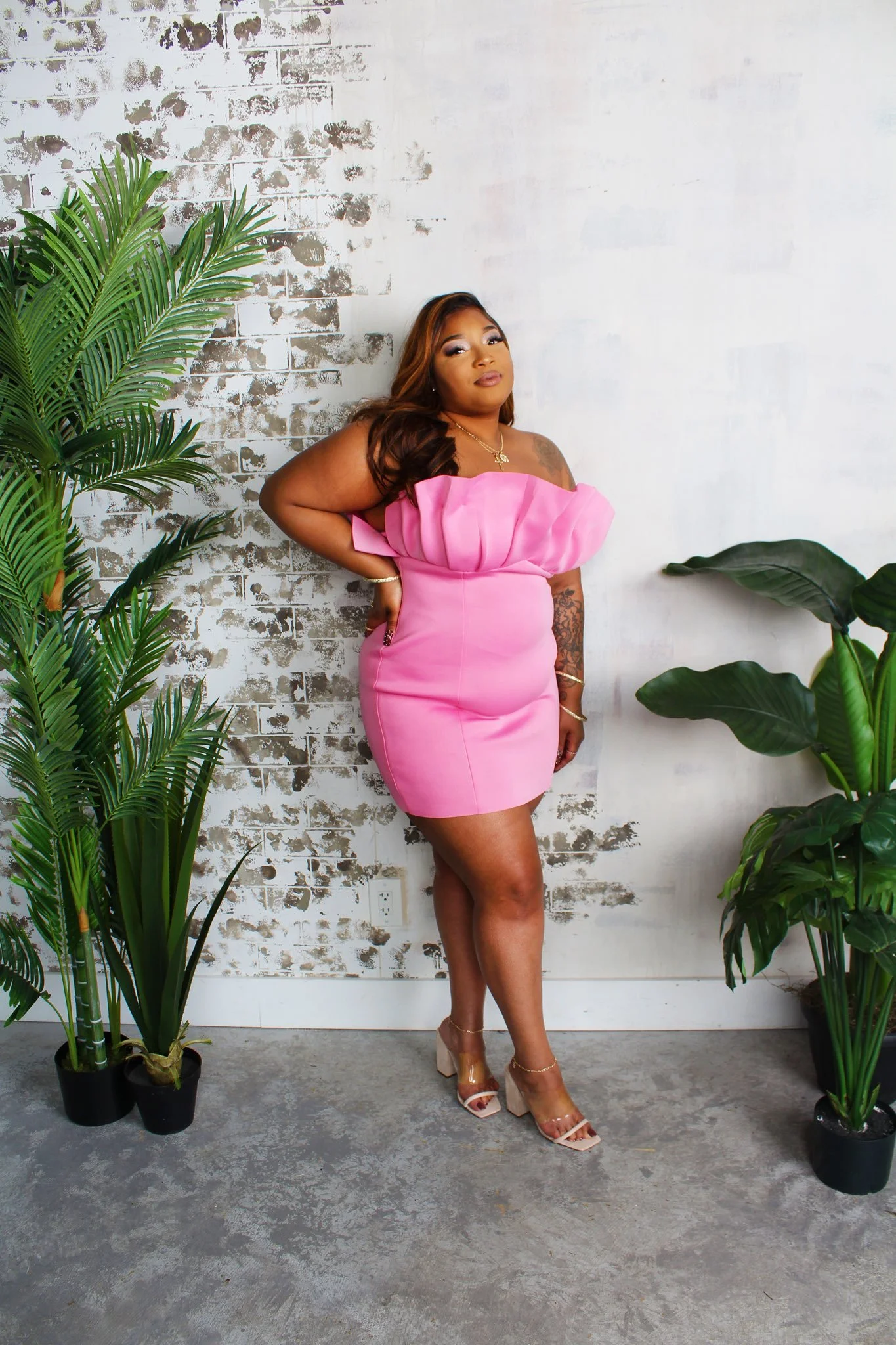 A woman in a bright pink dress posing indoors against a white wall with a black and white distressed pattern, flanked by two large green potted plants, wearing nude high heels and gold jewelry.