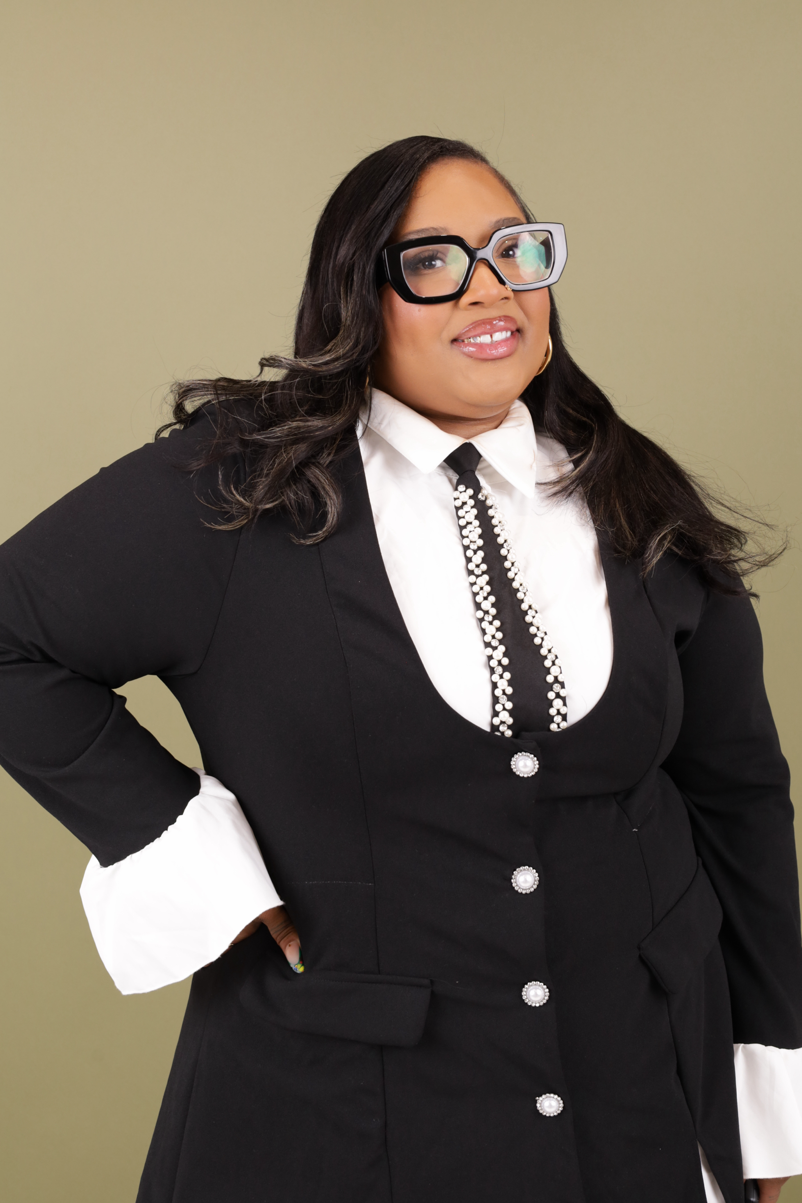 A woman with dark hair wearing large black glasses, a white collared shirt, a black jacket with pearl buttons, and a pearl necklace, standing against a plain background.