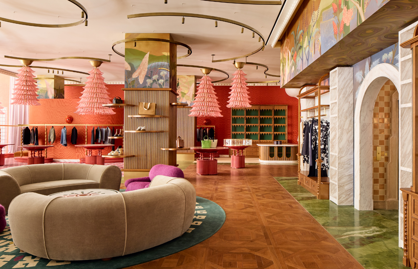 Interior of a colorful boutique with pink hanging lamps, red and green wall accents, display shelves with clothing and accessories, a curved beige sofa with purple chairs, and wooden flooring.