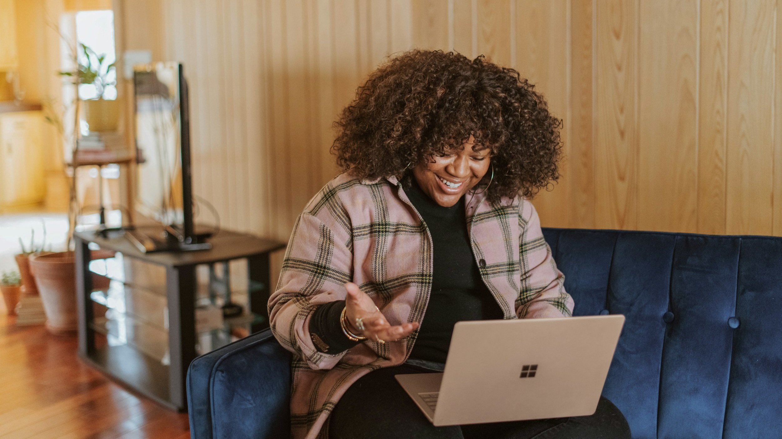 A woman with curly hair sits on a blue couch, smiling and gesturing while looking at a laptop in a cozy room with wooden walls.