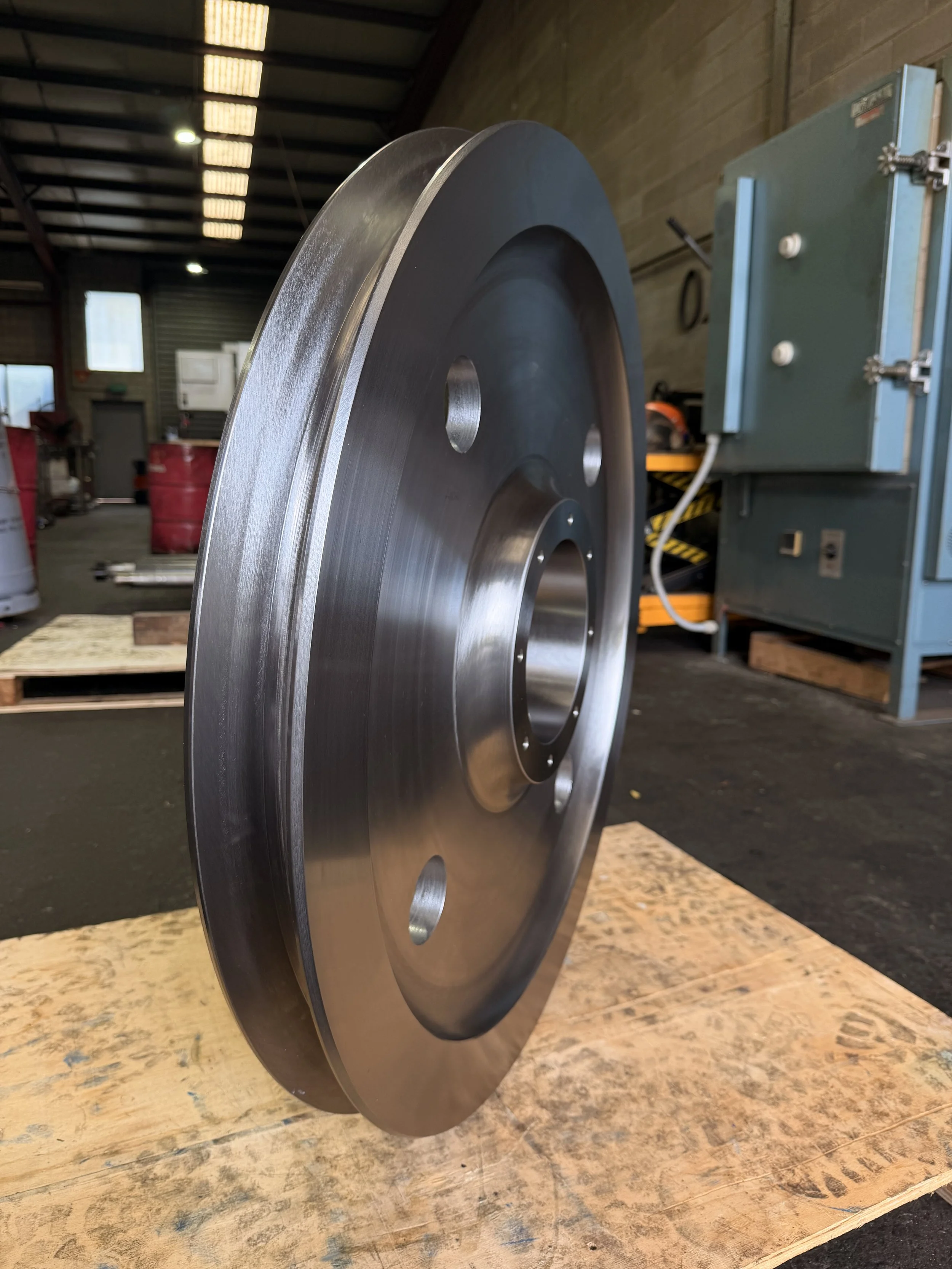 A large, shiny metal industrial wheel standing upright on a wooden surface inside a warehouse with a high ceiling and fluorescent lights.