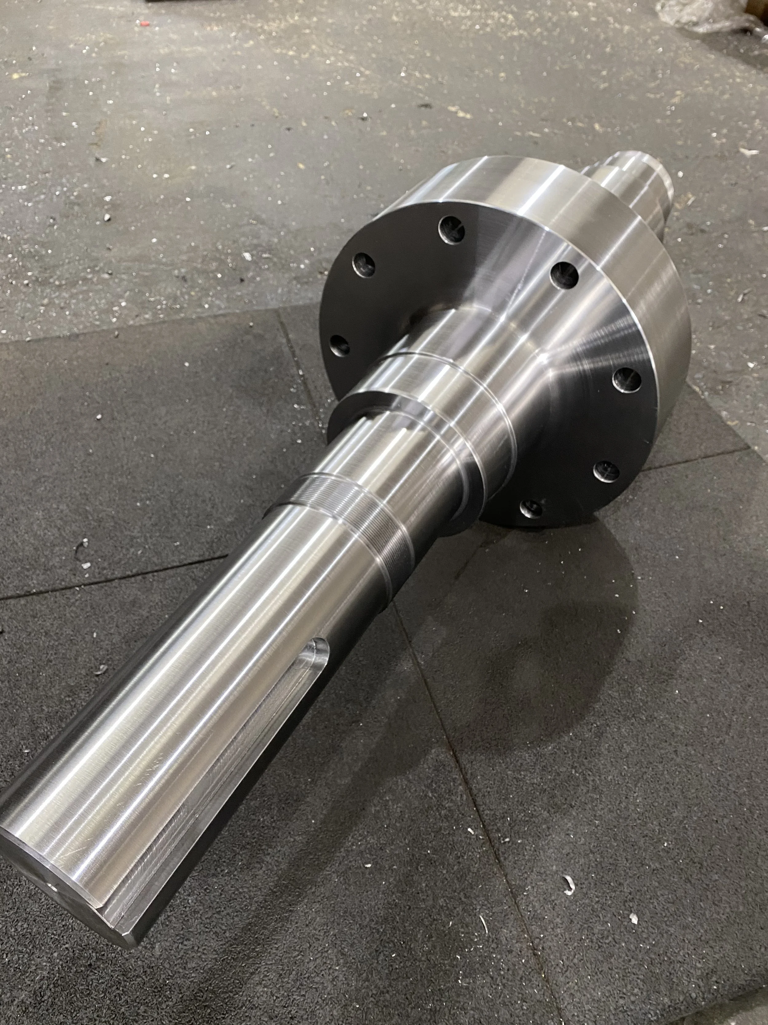 Large machined metal shaft resting on a dark surface, possibly in a workshop or manufacturing environment.