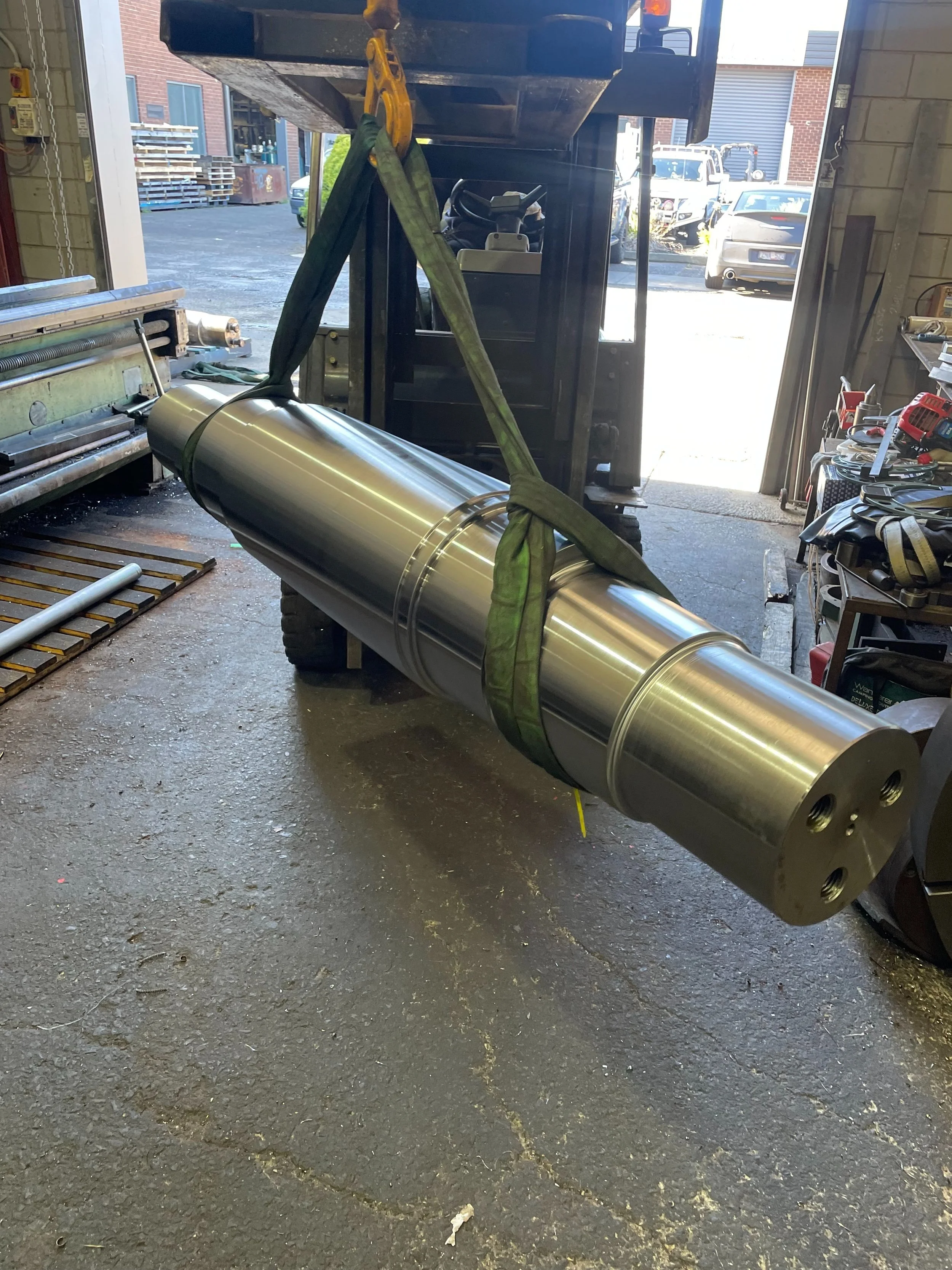 A forklift lifting a large, shiny, metallic cylindrical object inside a workshop. The workshop has various tools and equipment on shelves and tables, with an open garage door showing cars and a building outside.