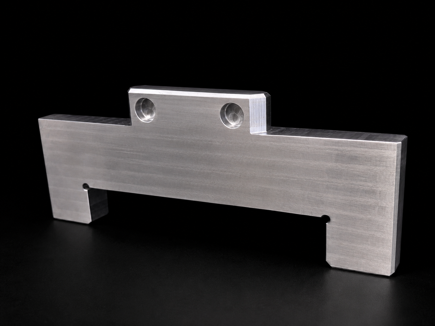 A metallic, rectangular machined part with two circular indentations and a protruding tab on one side, set against a black background.