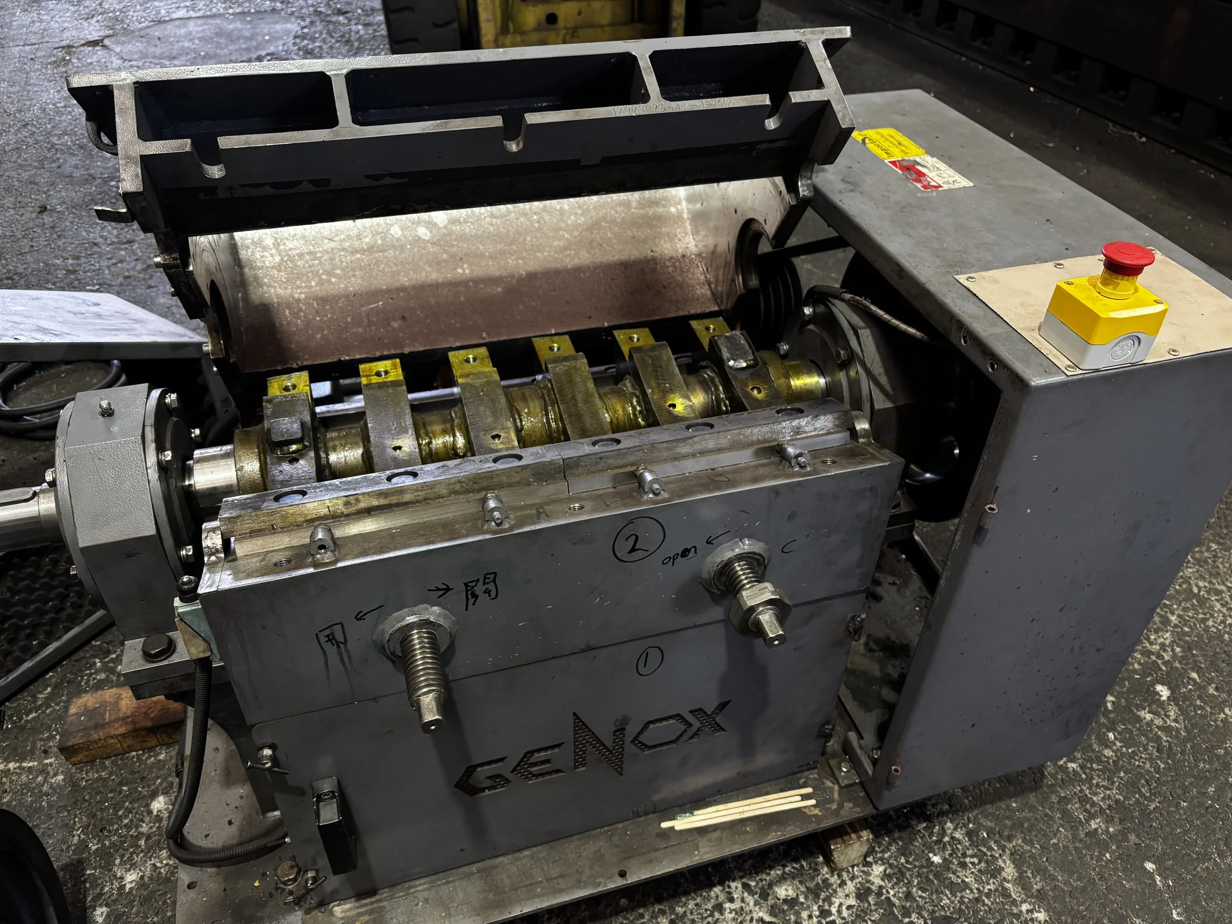 Close-up view of an industrial machine with gears, metal components, and a control box with a red emergency stop button in a workshop or factory setting.