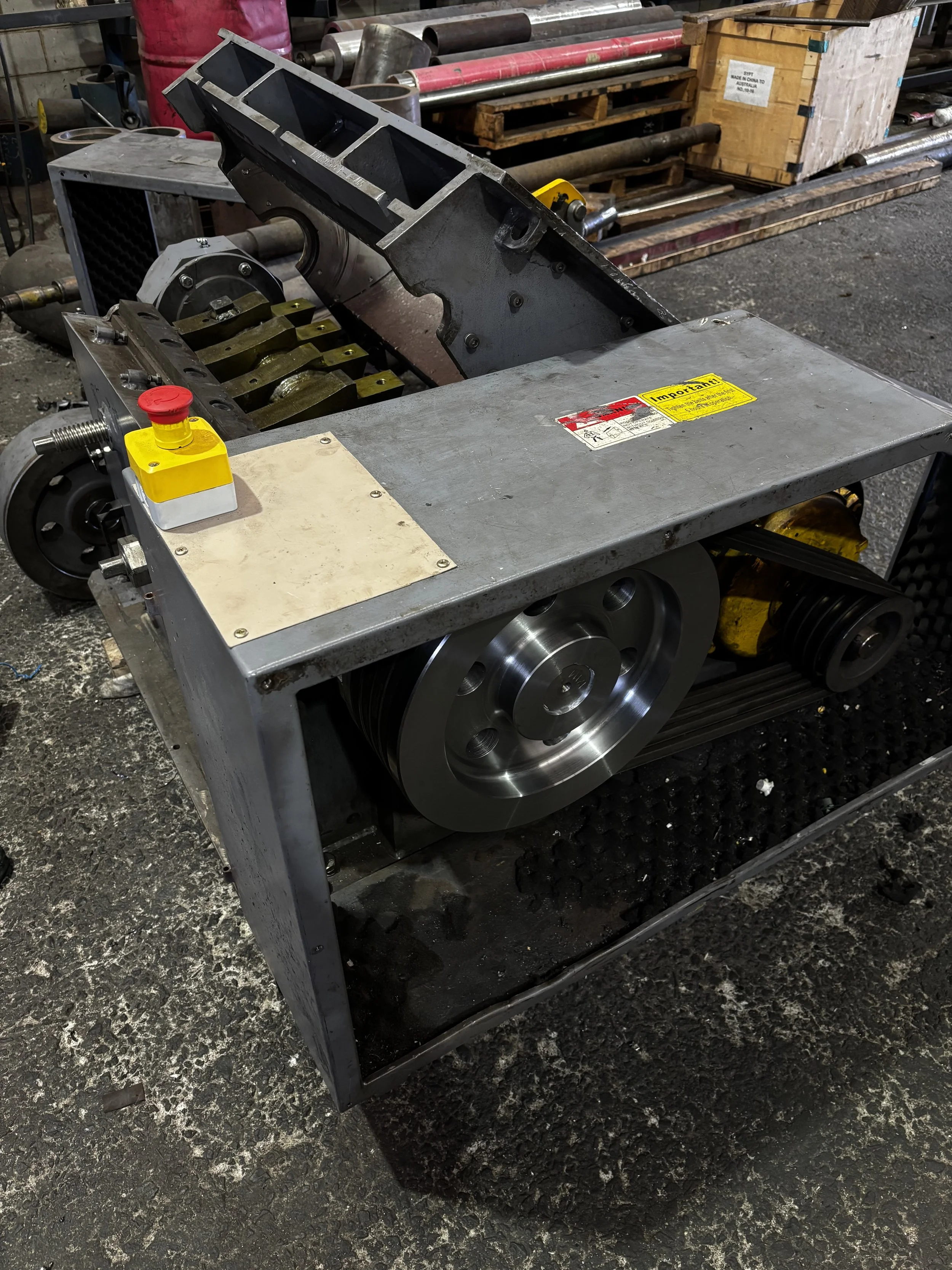 Photo of a mechanical assembly or equipment with metallic and industrial components in a workshop environment. The setup includes a large wheel, metal brackets, and a yellow warning label on a gray metal surface.