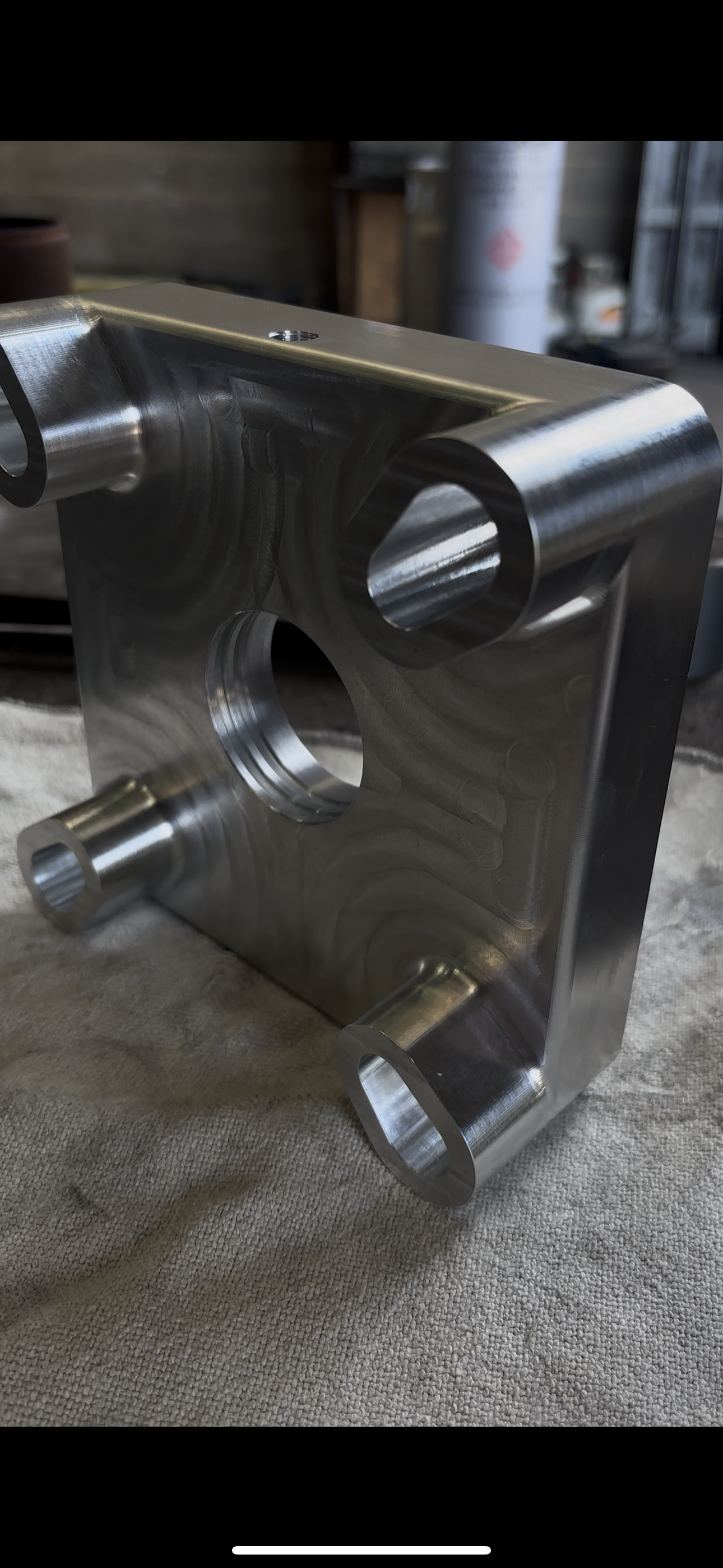 A machined metal block with four cylindrical protrusions and a central hole, sitting on a textured surface in a workshop setting.