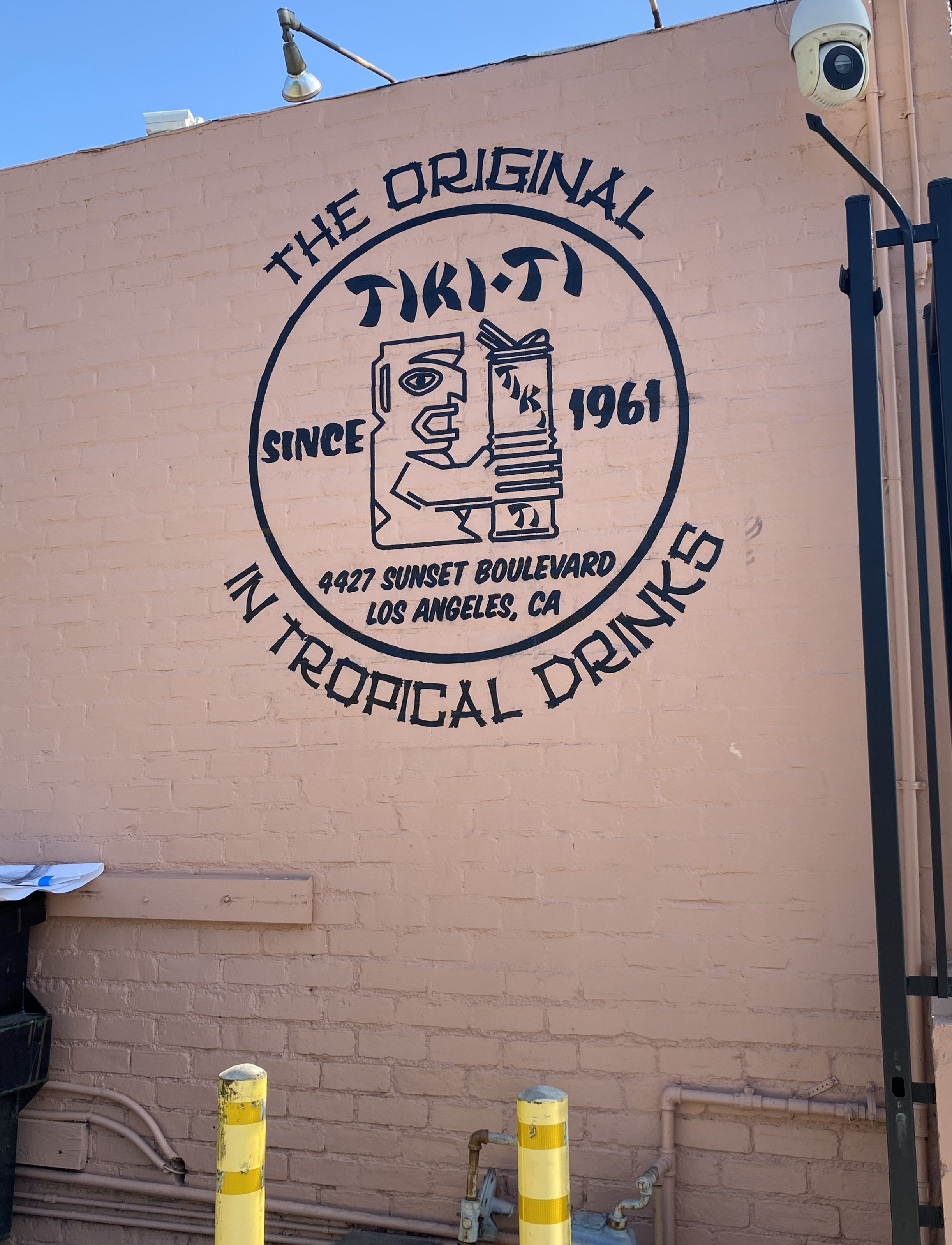 A mural on a beige brick wall advertising tiki ti, a bar established in 1961, located at 4427 Sunset Boulevard Los Angeles, CA, featuring a tiki idol holding a tiki drink, with the text 'The Original In Tropical Drinks'.