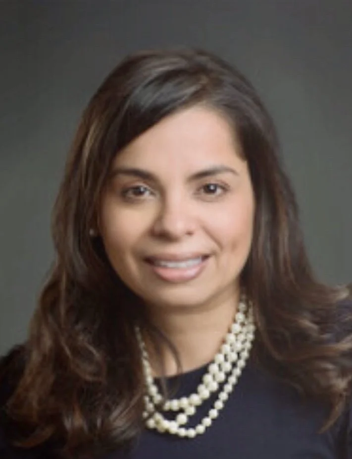 A woman with long, dark, wavy hair wearing a black top and a string of pearls, smiling at the camera against a plain background.