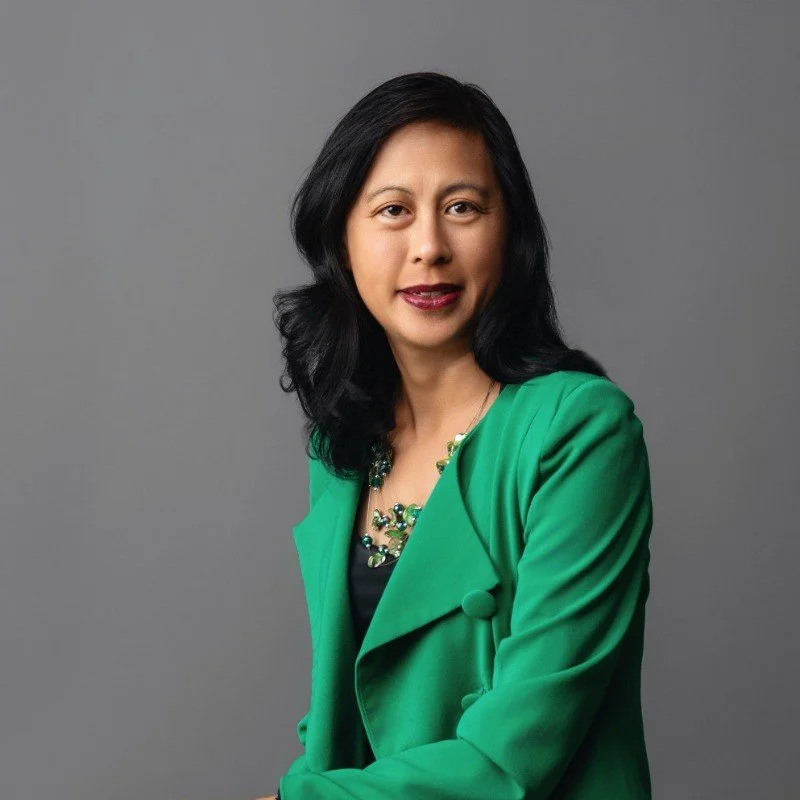 A woman with black hair wearing a green blazer and a colorful necklace, posing against a gray background.