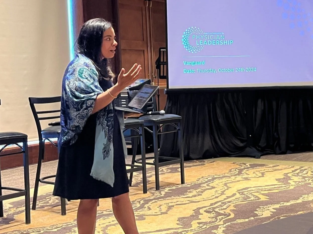 A woman giving a presentation at a conference, standing near a large screen displaying a slide titled 'American Association for the Physician Leadership' with details about the event and date, while gesturing with her hand.