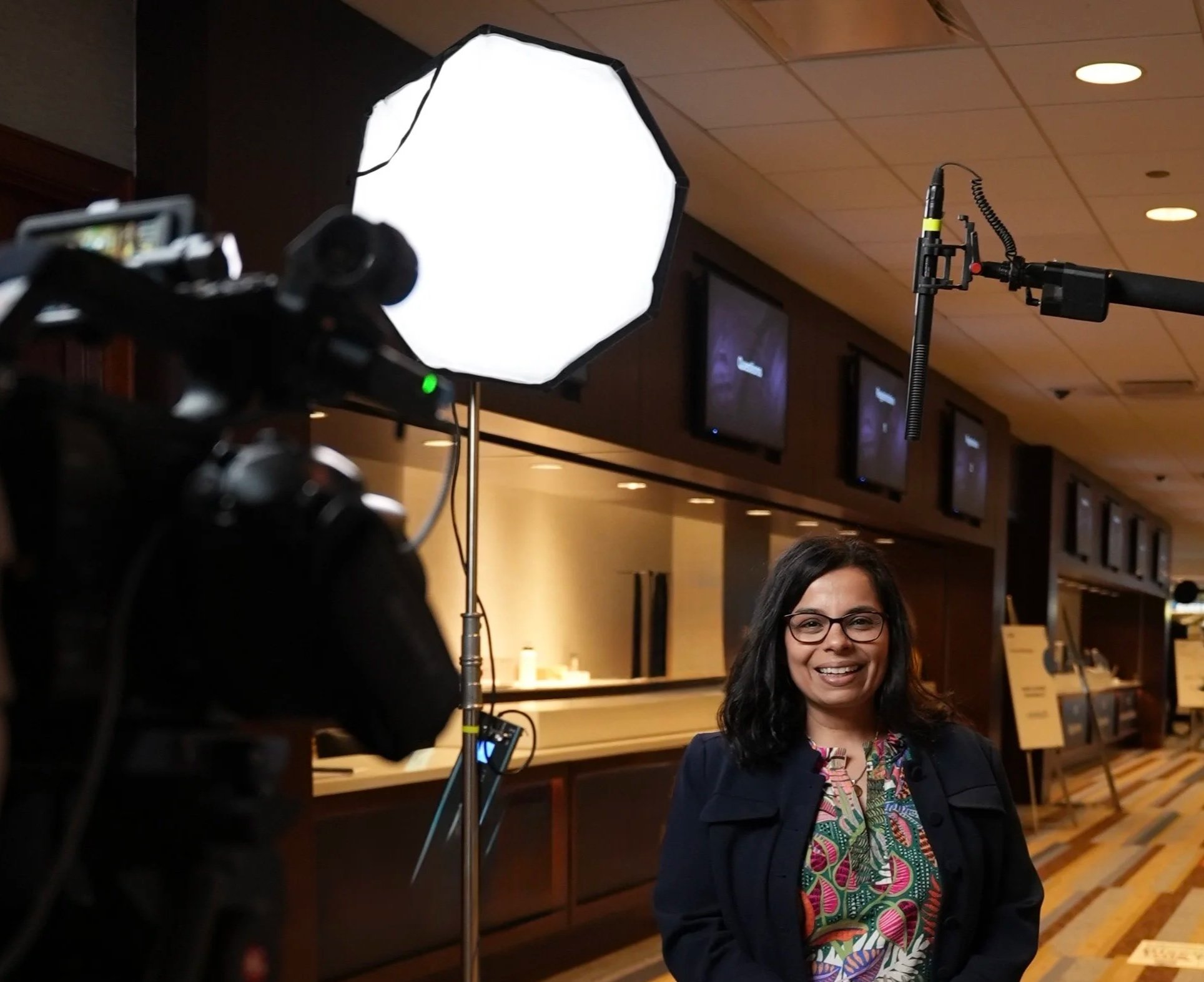 A woman with dark hair and glasses smiling at camera in a media interview setting, with studio lights and camera equipment visible.
