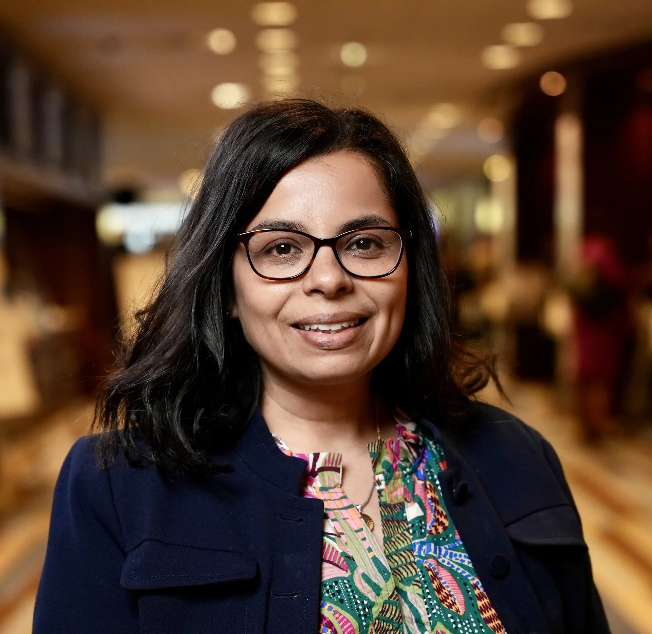 A woman with black hair and glasses smiling in a warmly lit indoor setting.